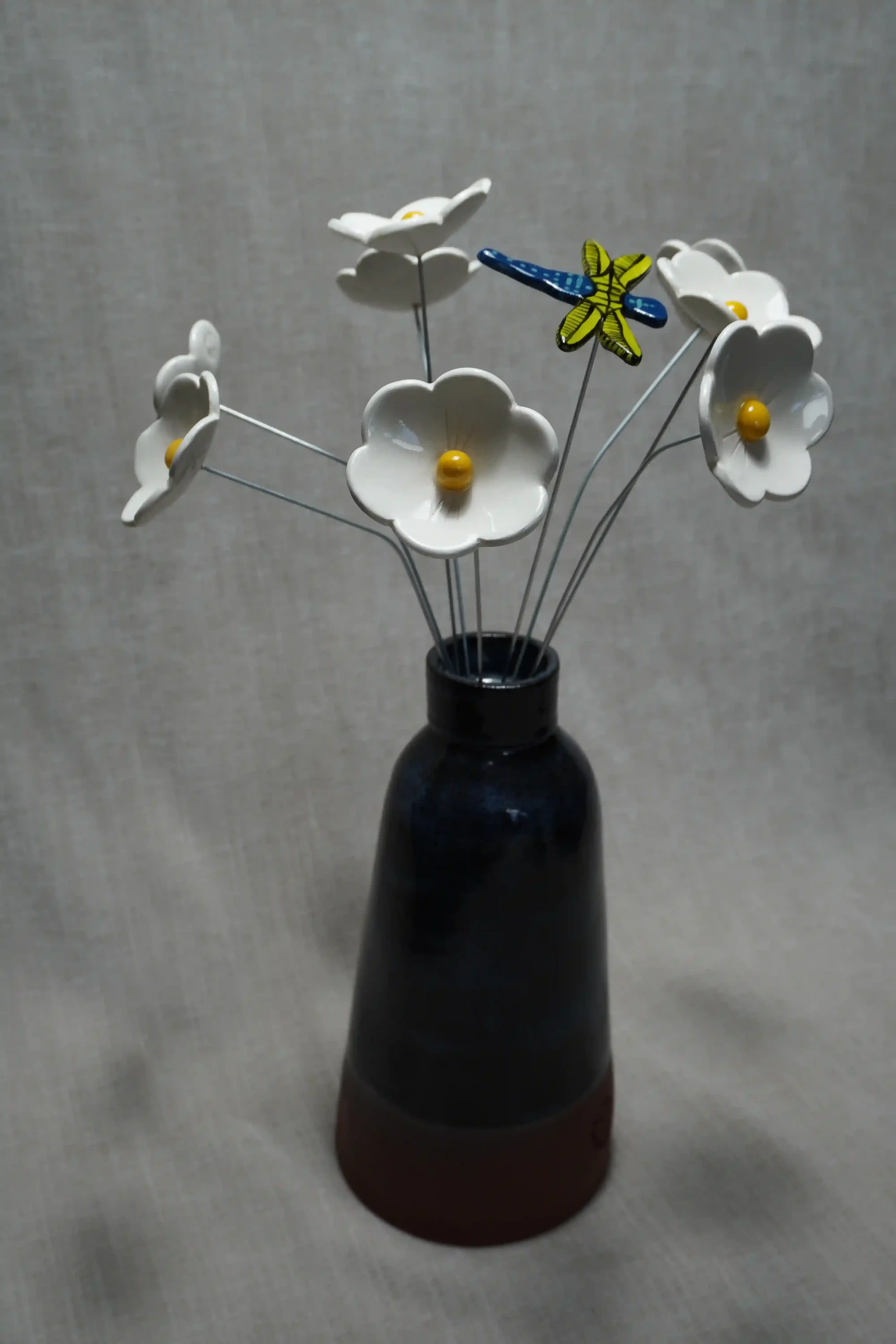 A dark blue and terracotta ceramic vase holding ceramic white Daisy flowers with yellow centers, and a colorful ceramic dragonfly with turquoise body with blue spots, black and light blue wings, on thin metal stems.