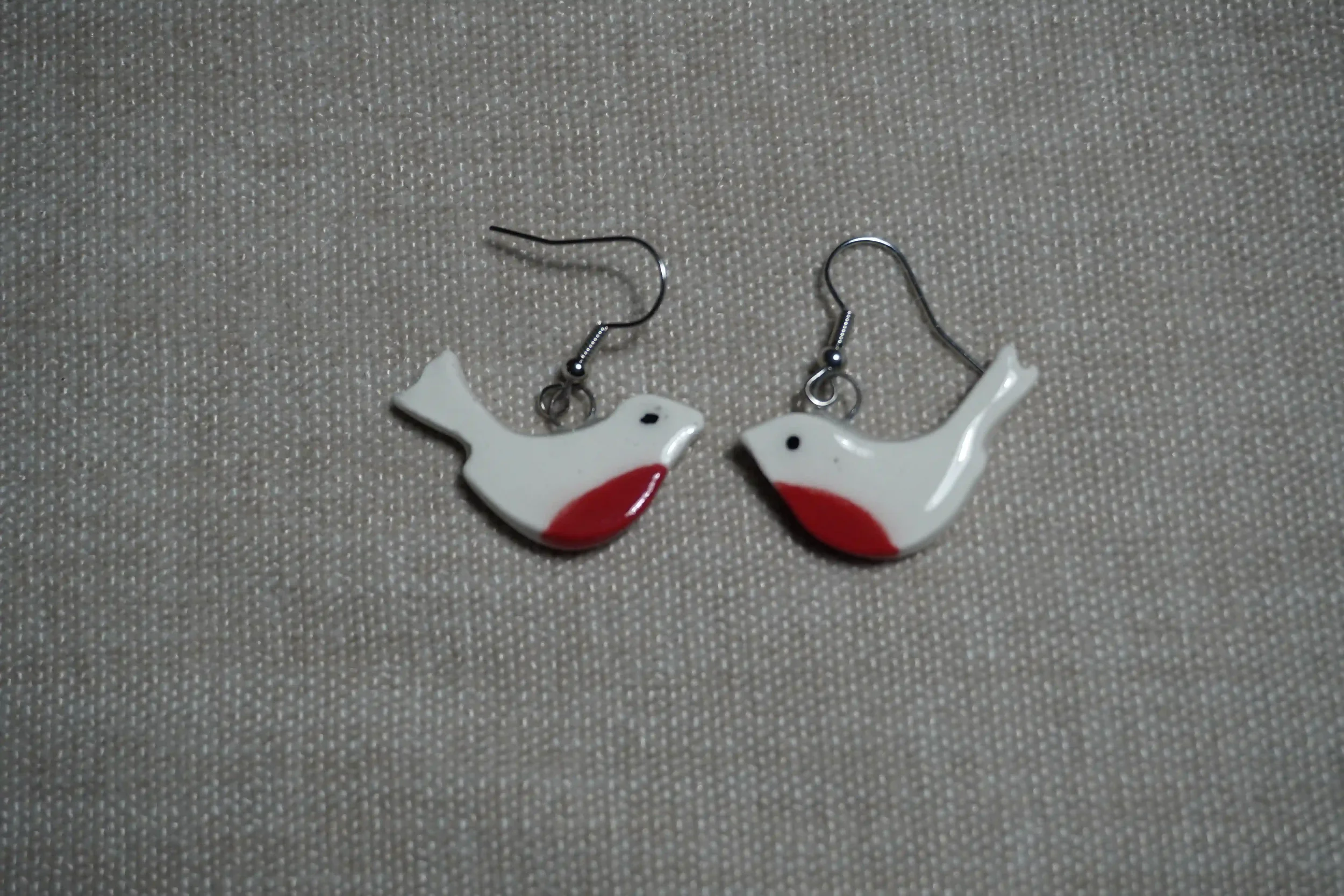 Pair of ceramic bird-shaped earrings with white bodies, red breast, and black eyes, placed on a textured neutral surface.
