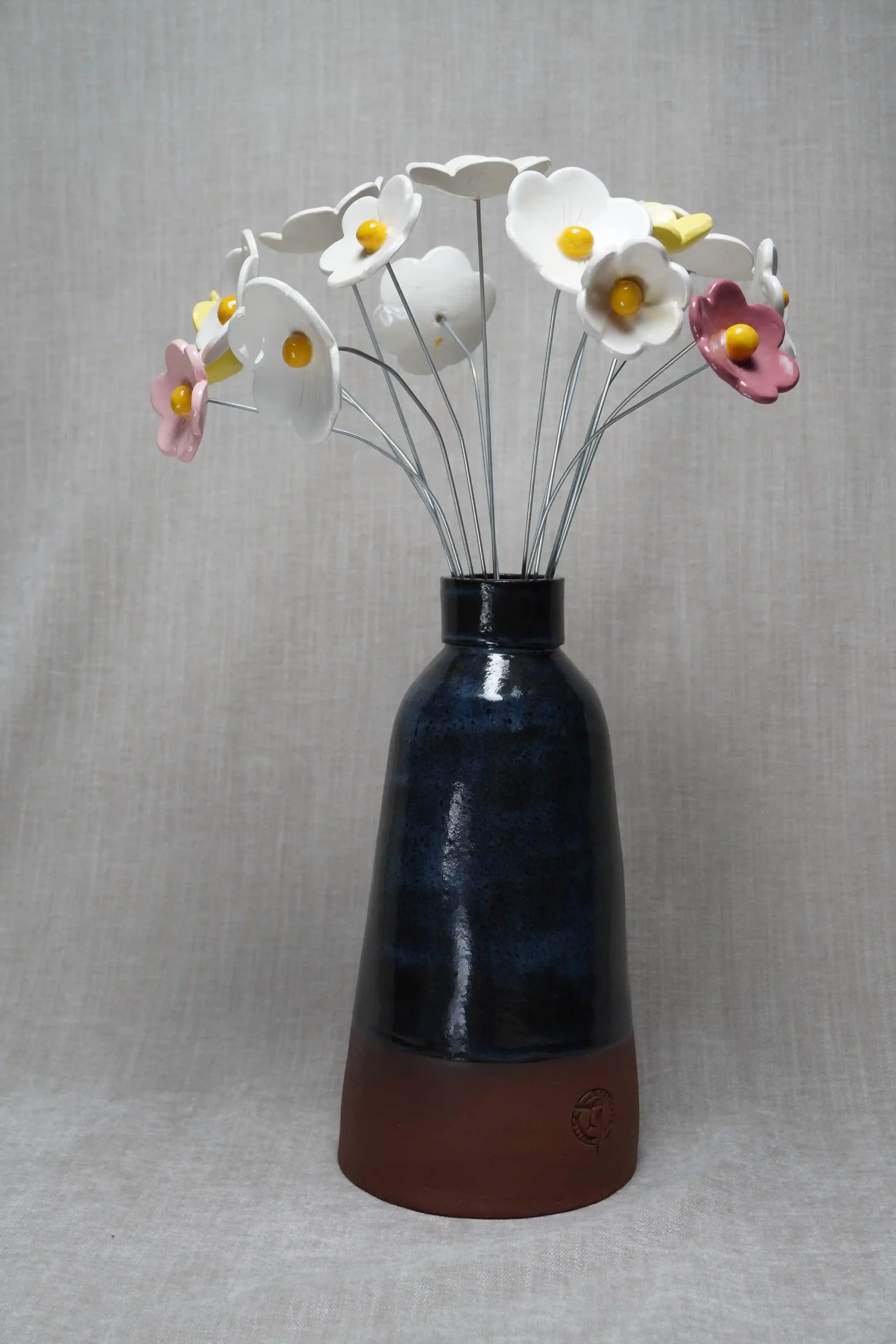 A ceramic pottery vase with a wide, rounded terracotta base, narrow neck, and a blue glaze, holding thirteen ceramic, pink, yellow and white flowers with yellow centres and metal stems, displayed on a light neutral background.