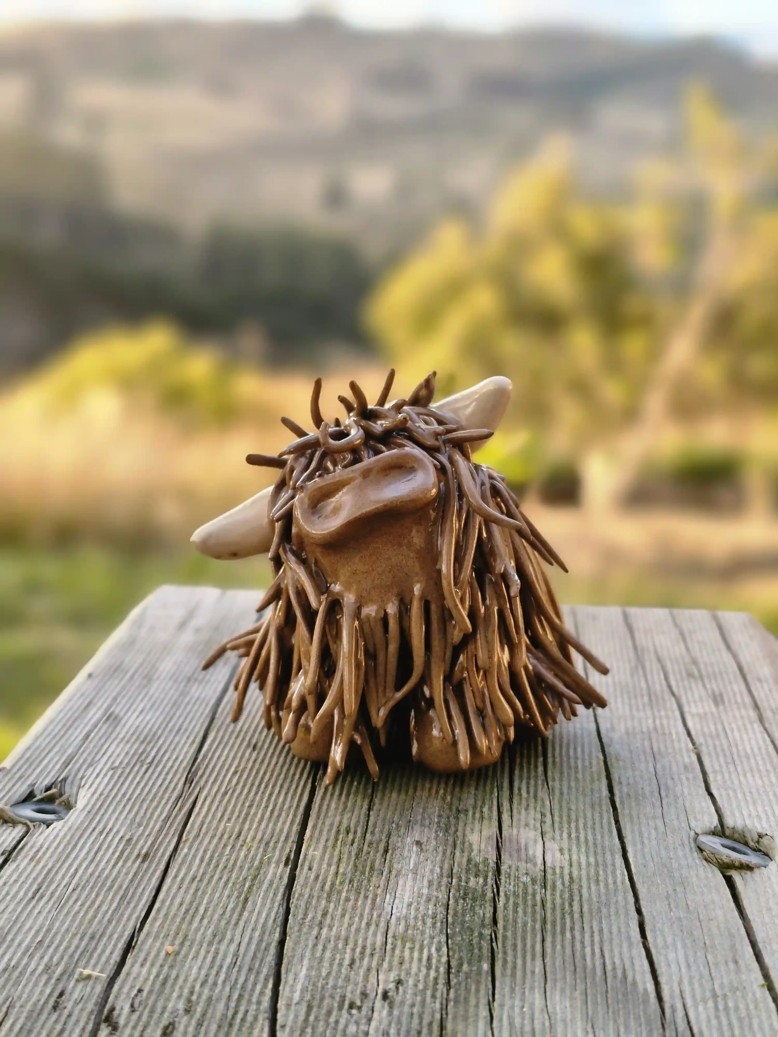 A small ceramic pottery figurine of a cute HIGHLAND COW standing on a weathered wooden surface outdoors with a blurred landscape background.