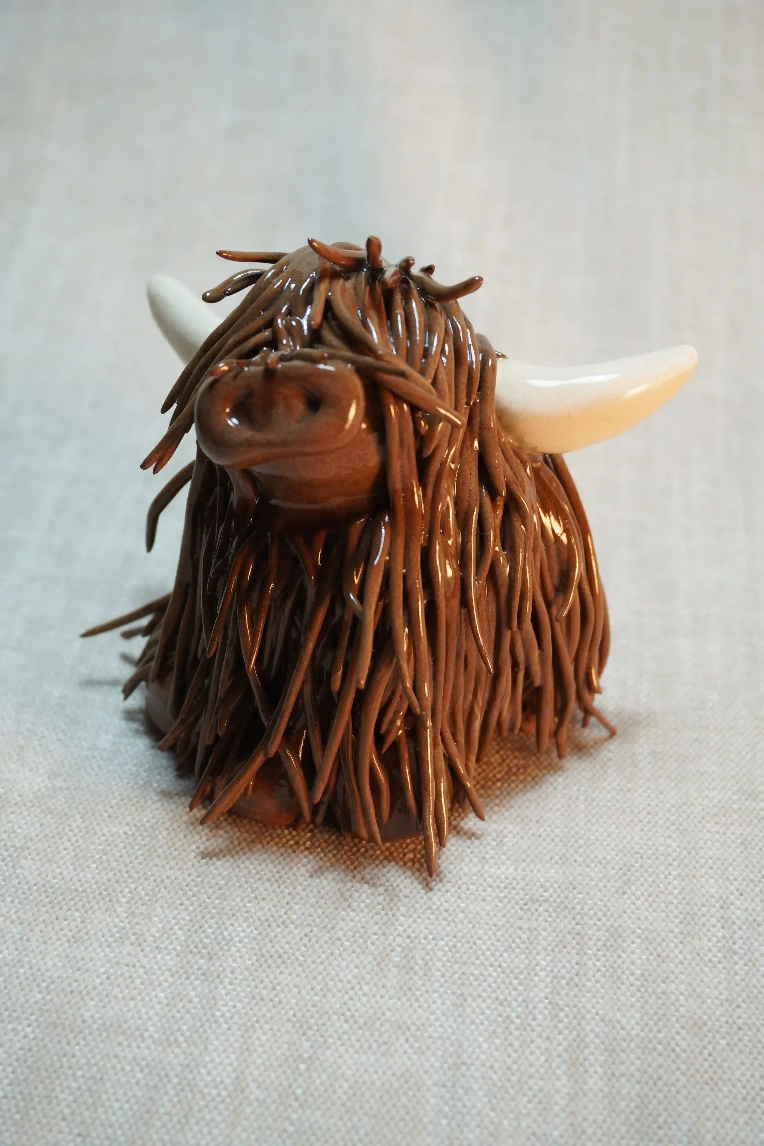 Front view of a handmade ceramic sculpture of a Highland Cow ornament with rich dark brown-coloured hair and beige horns.