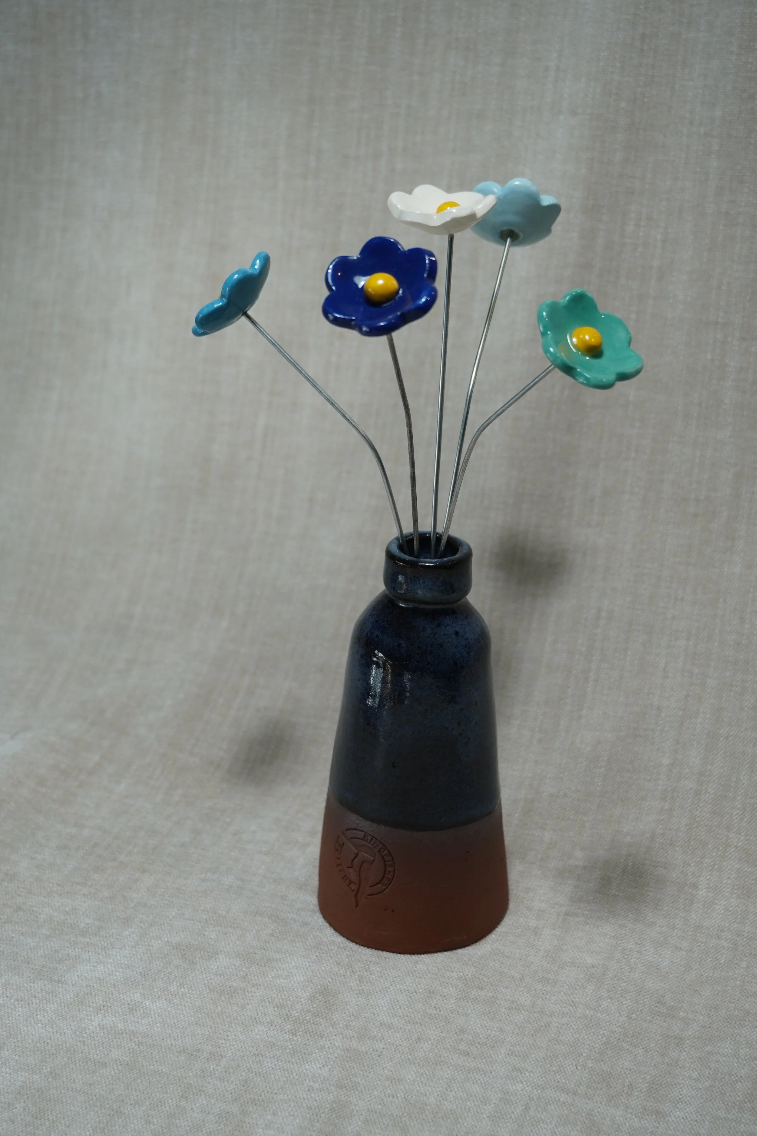 A ceramic vase with a wide, rounded terracotta base, narrow neck, and a blue glaze,  holding five ceramic flowers in white, dark blue, light blue, green and teal with yellow centers and metal stems, standing on a neutral textured fabric surface.