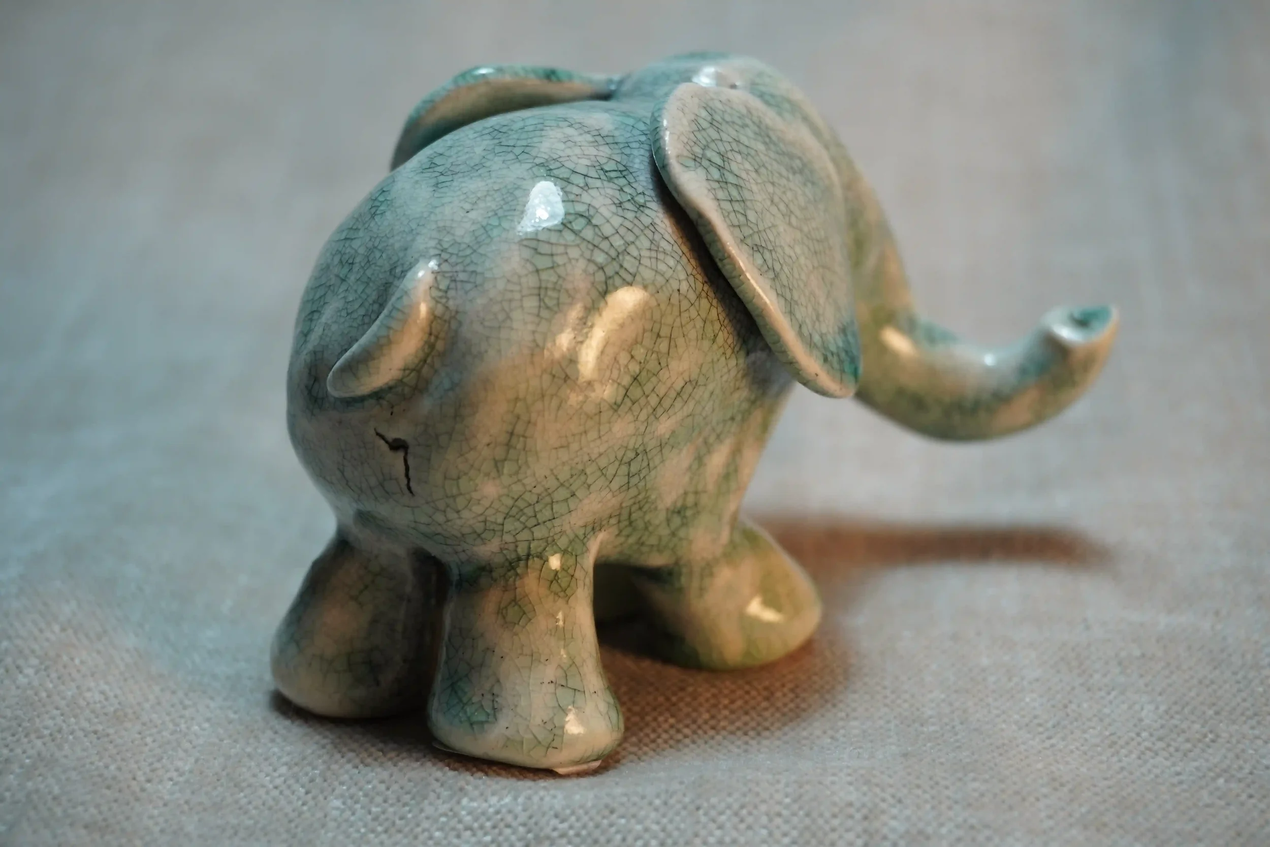 Rear view of a small ceramic figurine of an elephant with a textured, blue-green crackled glaze finish, placed on a neutral surface.