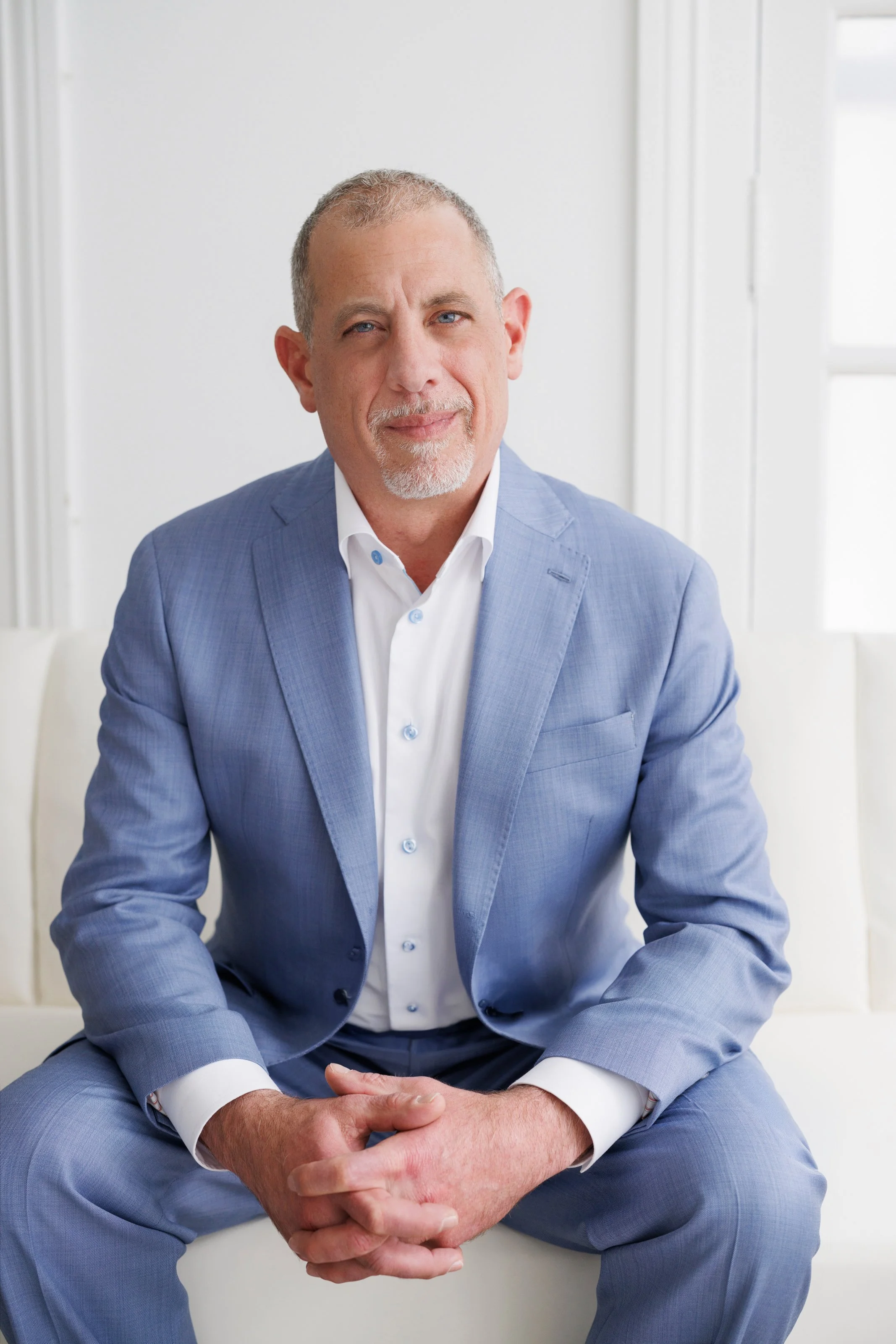 A middle-aged man with short gray hair and a beard, wearing a light blue suit and white shirt, sitting with hands clasped and looking at the camera in a well-lit room with white walls and a window.