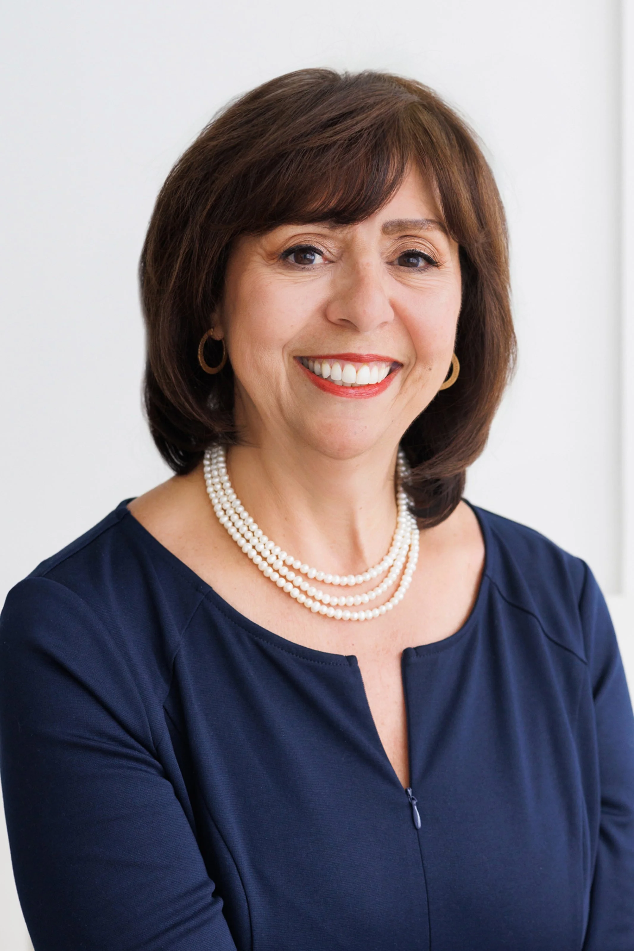 A woman with short brown hair smiling, wearing a navy blue top, pearl necklace, and hoop earrings, in a professional setting.