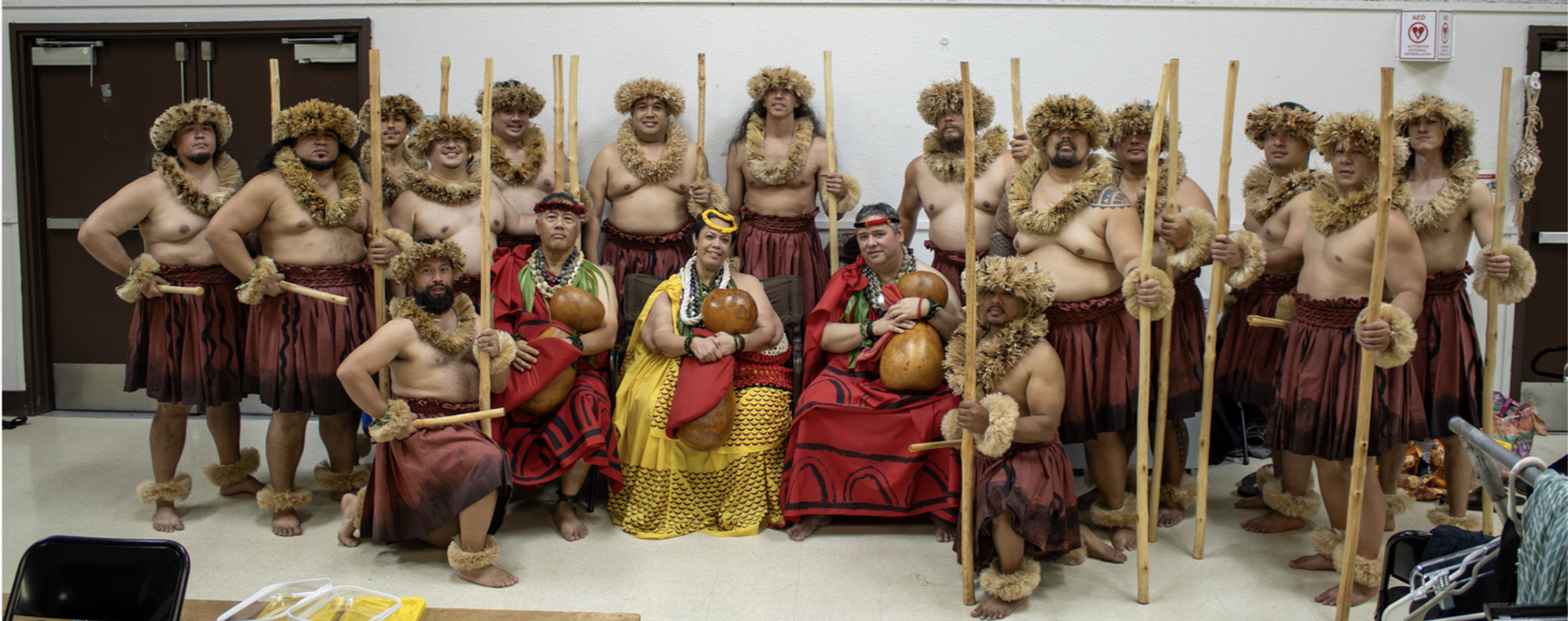 Group of men and women dressed in traditional Polynesian costumes, holding wooden staffs and gourds, posing indoors against a white wall.