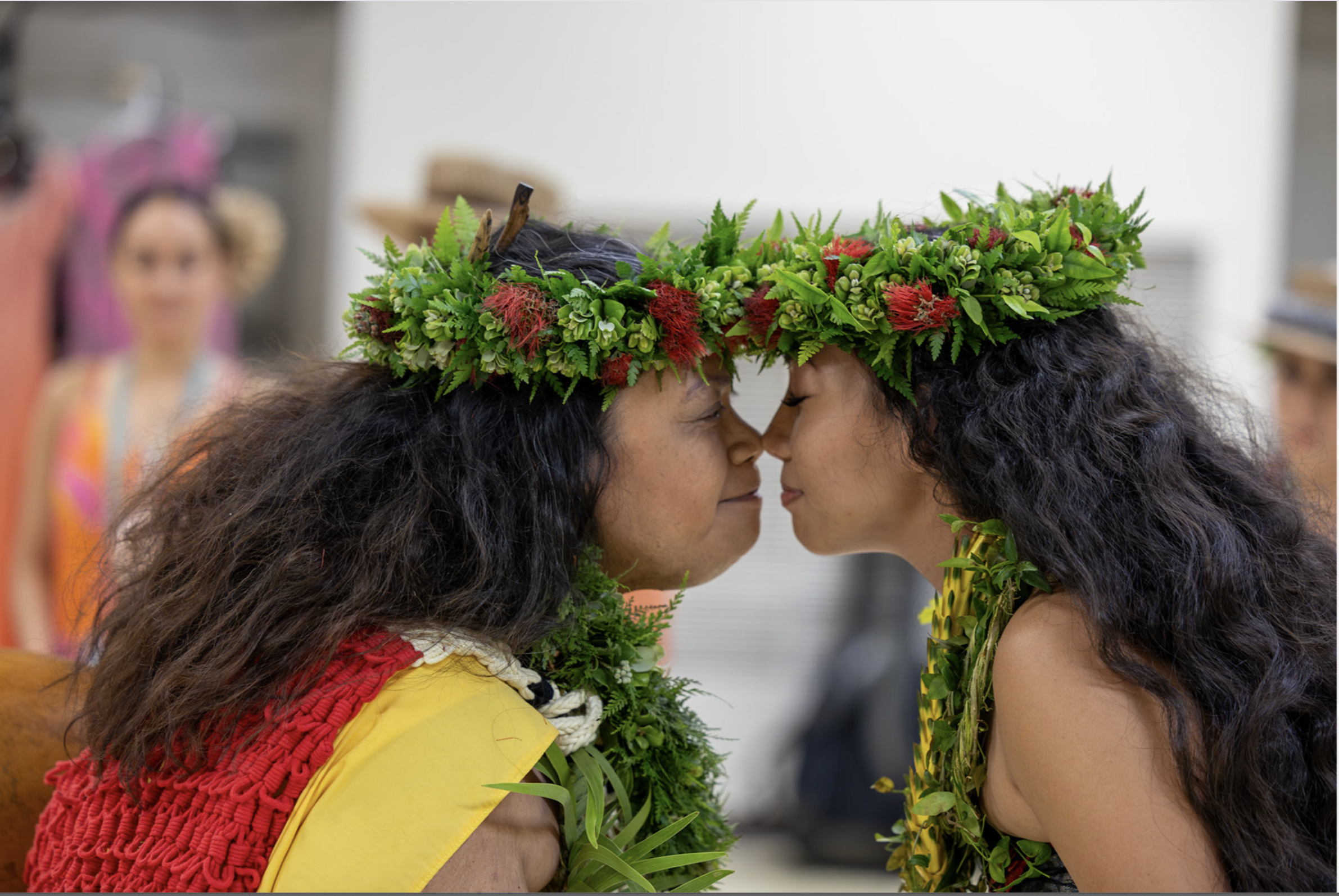 Two women in traditional attire face each other, wearing crowns made of lush green leaves and red flowers, with their foreheads touching.