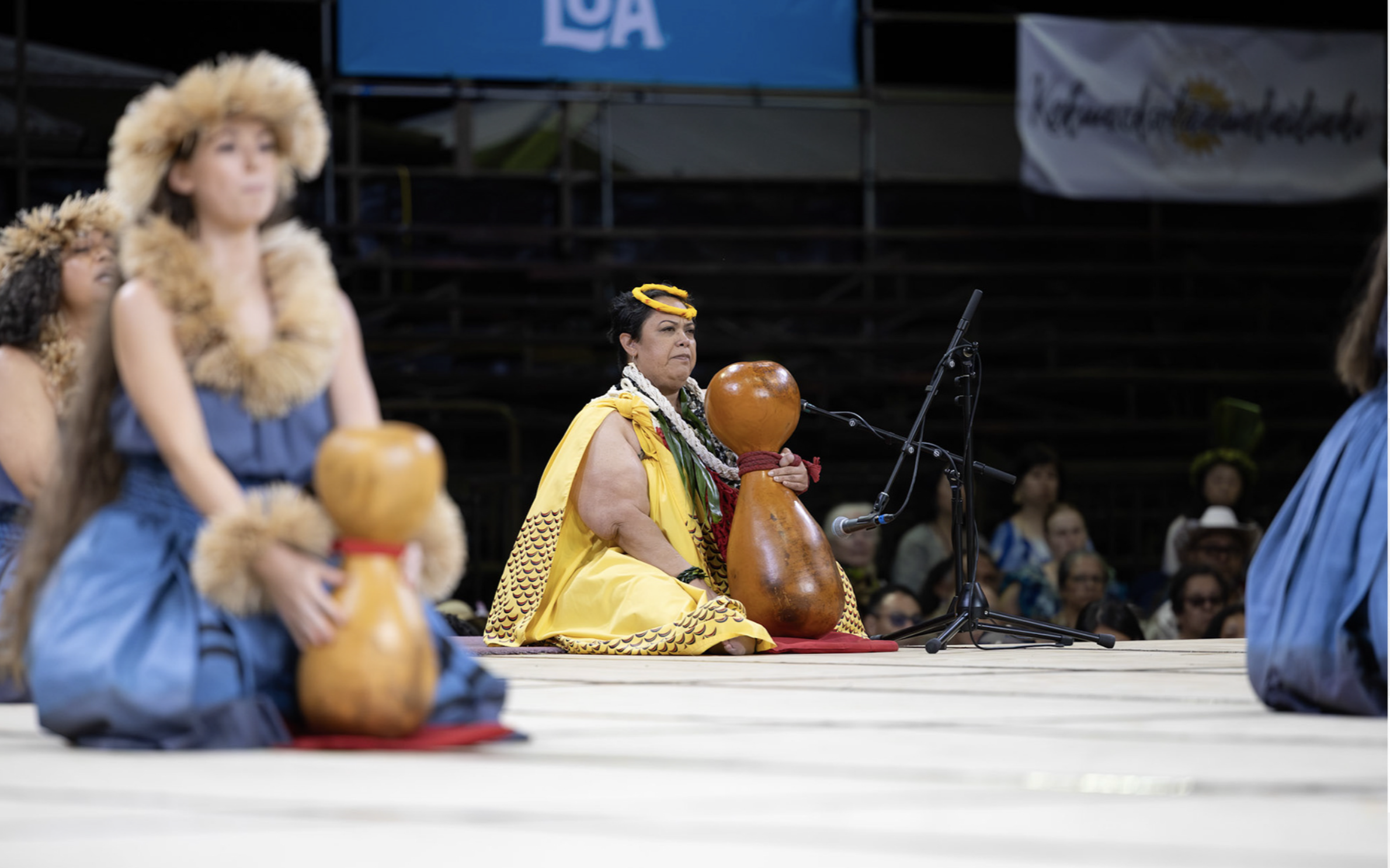 Native Hawaiian performers sit on stage, wearing traditional clothing and accessories, with a woman playing a large gourd drum in the center. Audience members are visible in the background.