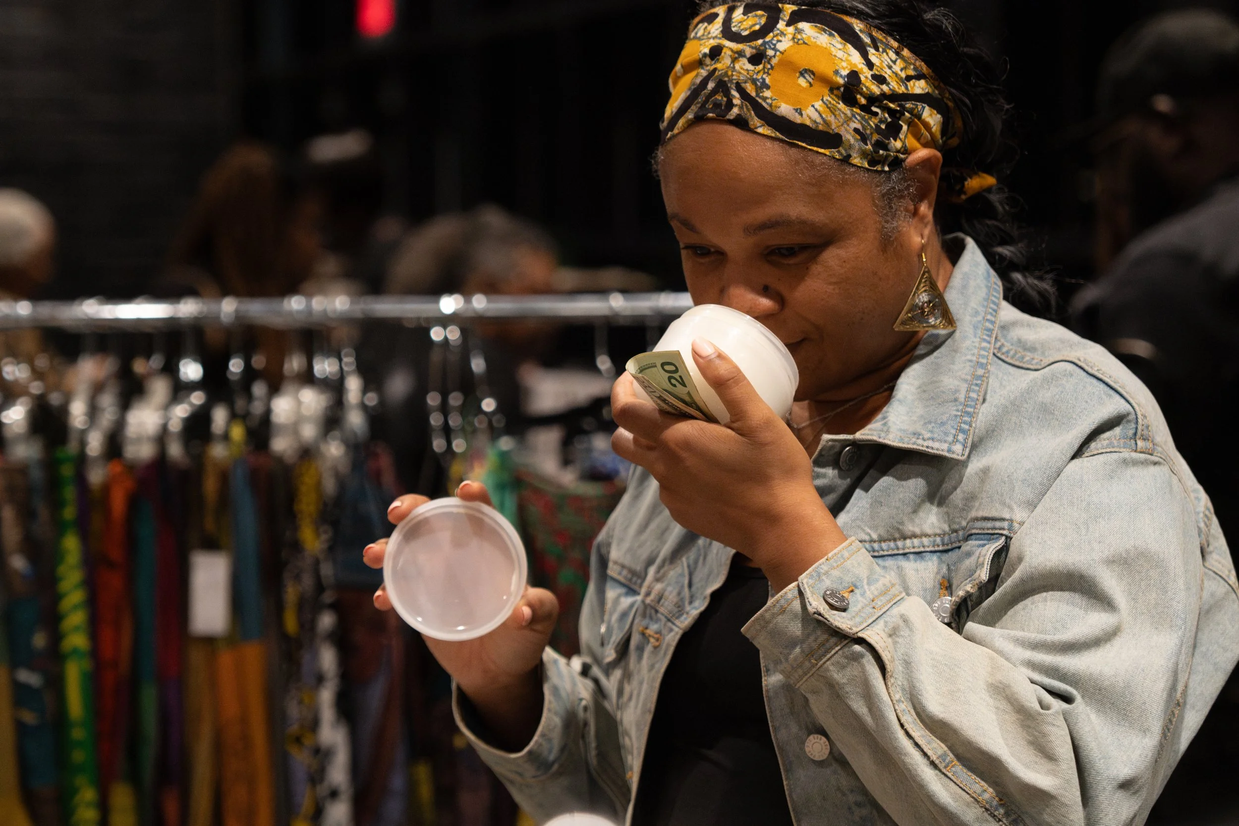 A woman wearing a yellow patterned headscarf and denim jacket is holding a white container with a twenty-dollar bill inside, looking at it inside a store, with various products in the background.