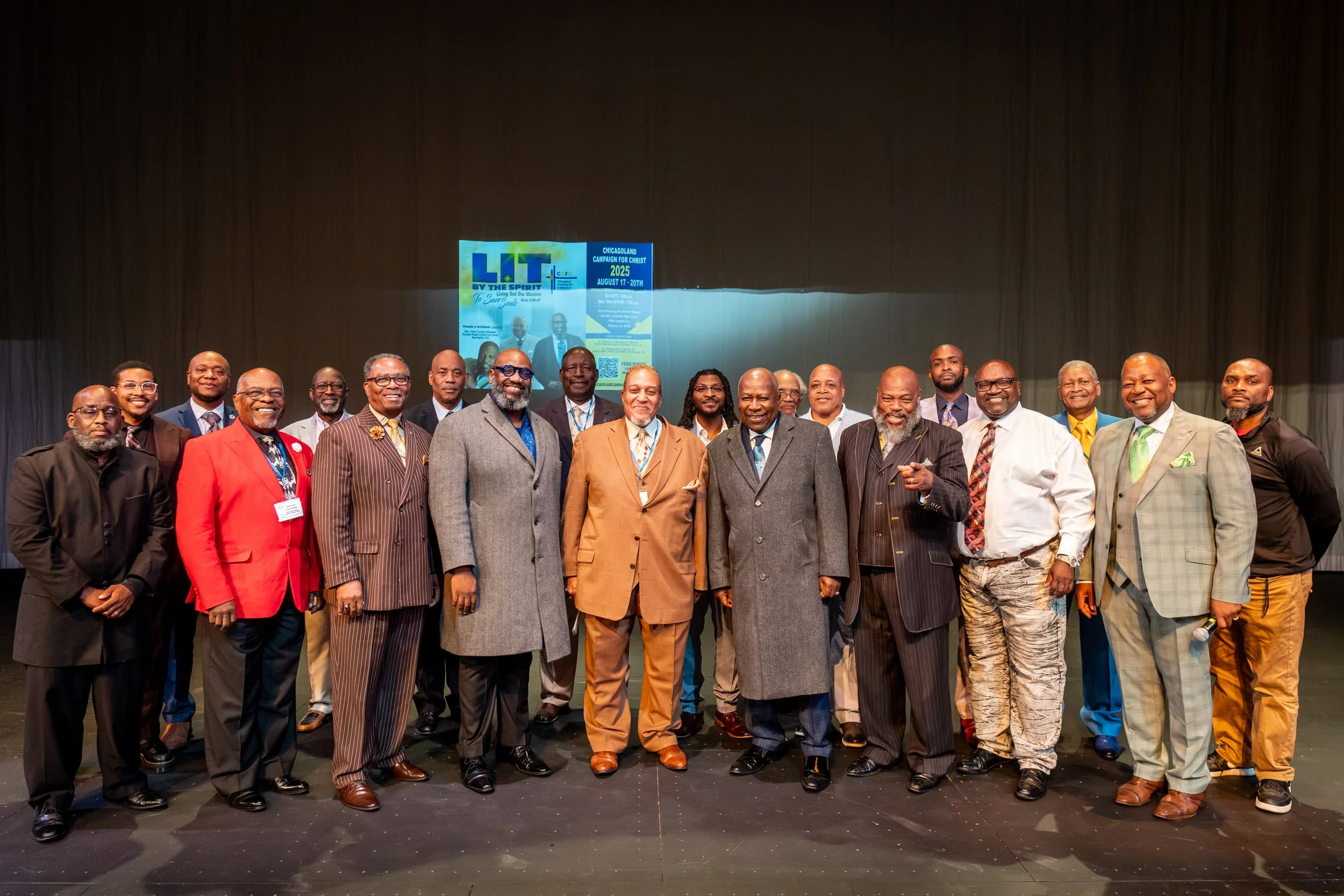 A group of men in formal and casual attire posing on stage in front of a screen displaying a flyer for a religious event in August 2025.