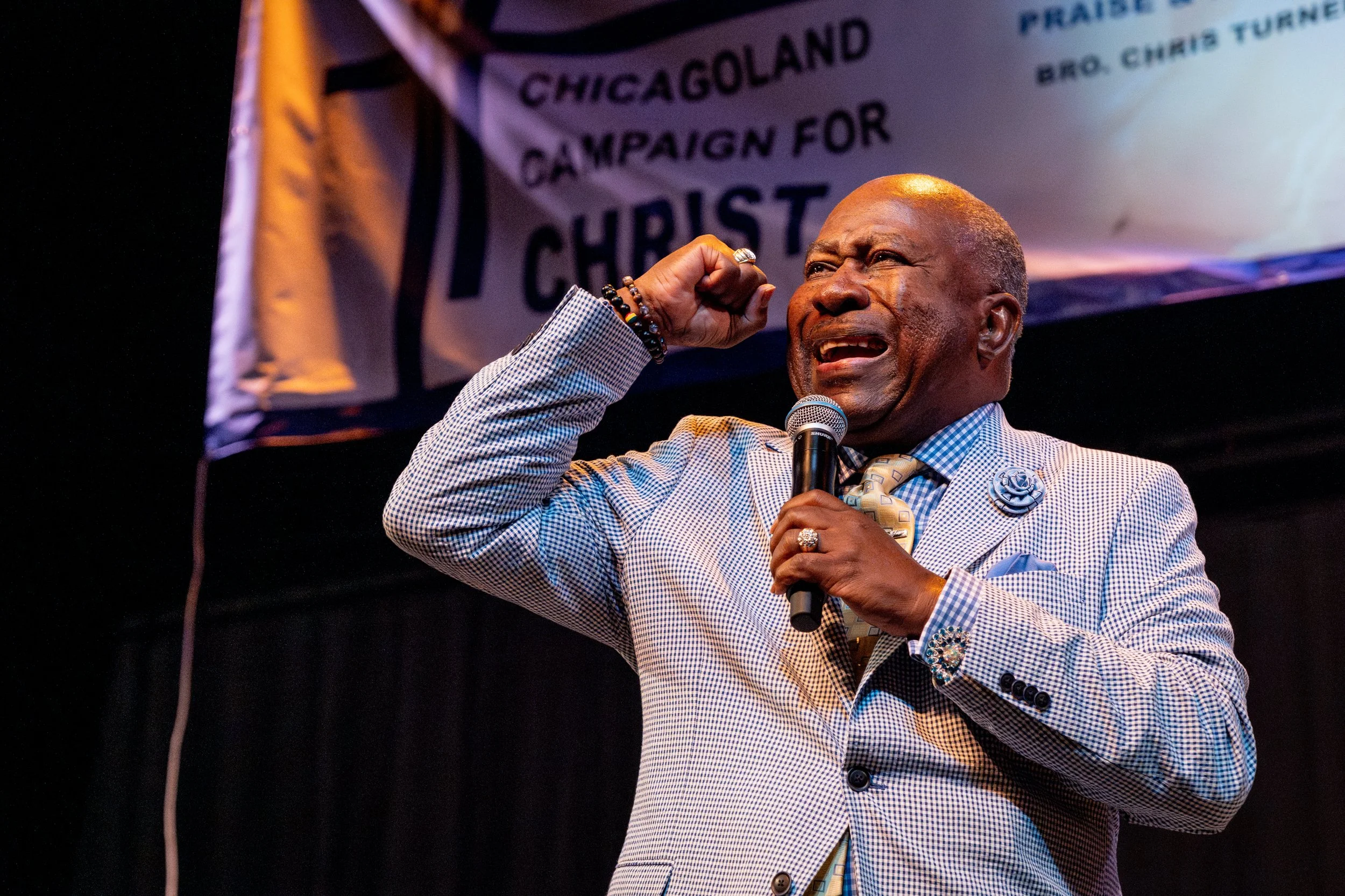 An elderly man with gold hair and dark skin passionately speaking into a microphone while raising his right fist. He is dressed in a checked blazer, a light-colored shirt, and a light tie, accessorized with rings, bracelets, and a lapel pin. Behind h