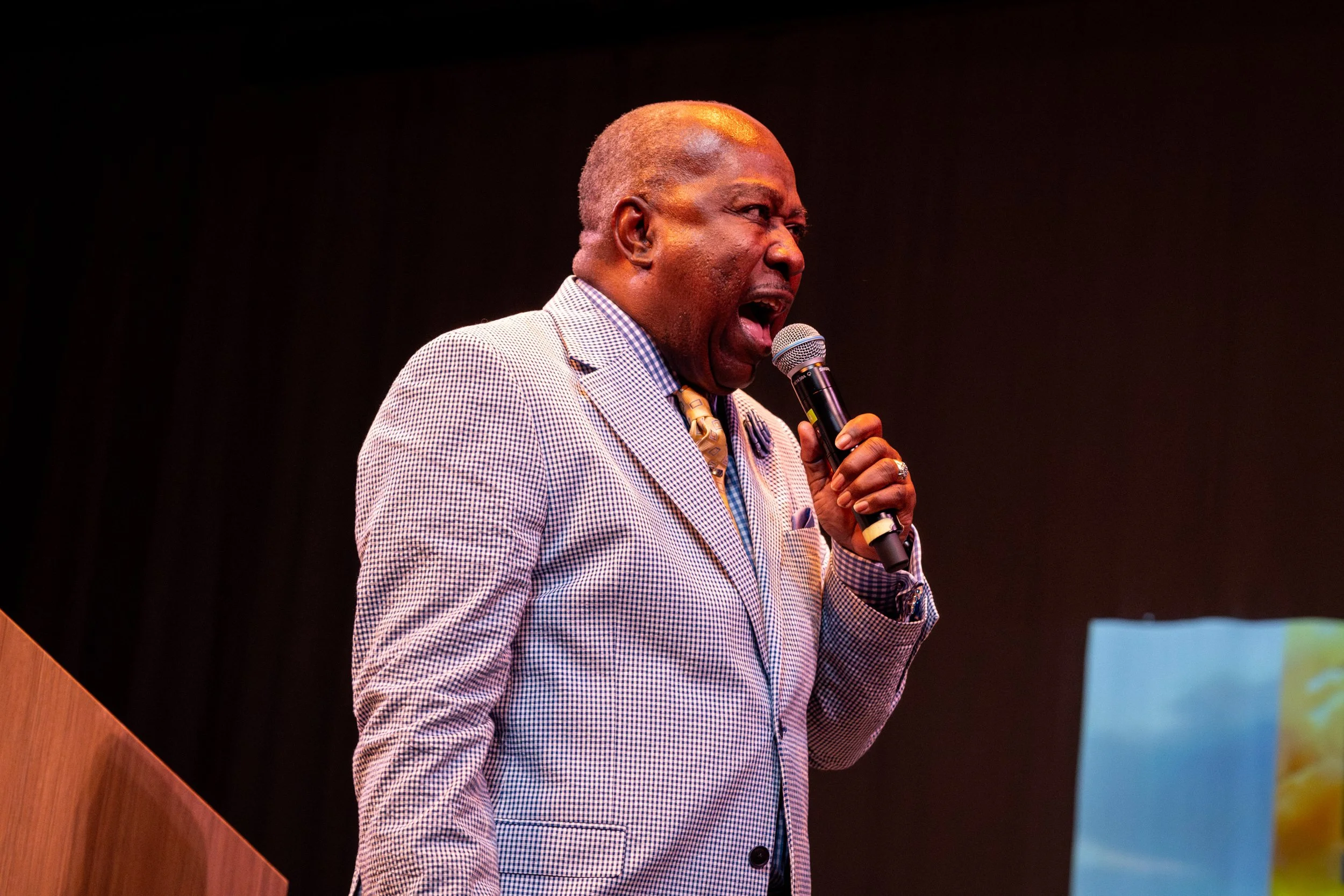 A man with dark skin and short gray hair speaking into a microphone at a presentation or event, dressed in a checked suit and standing next to a wooden desk.