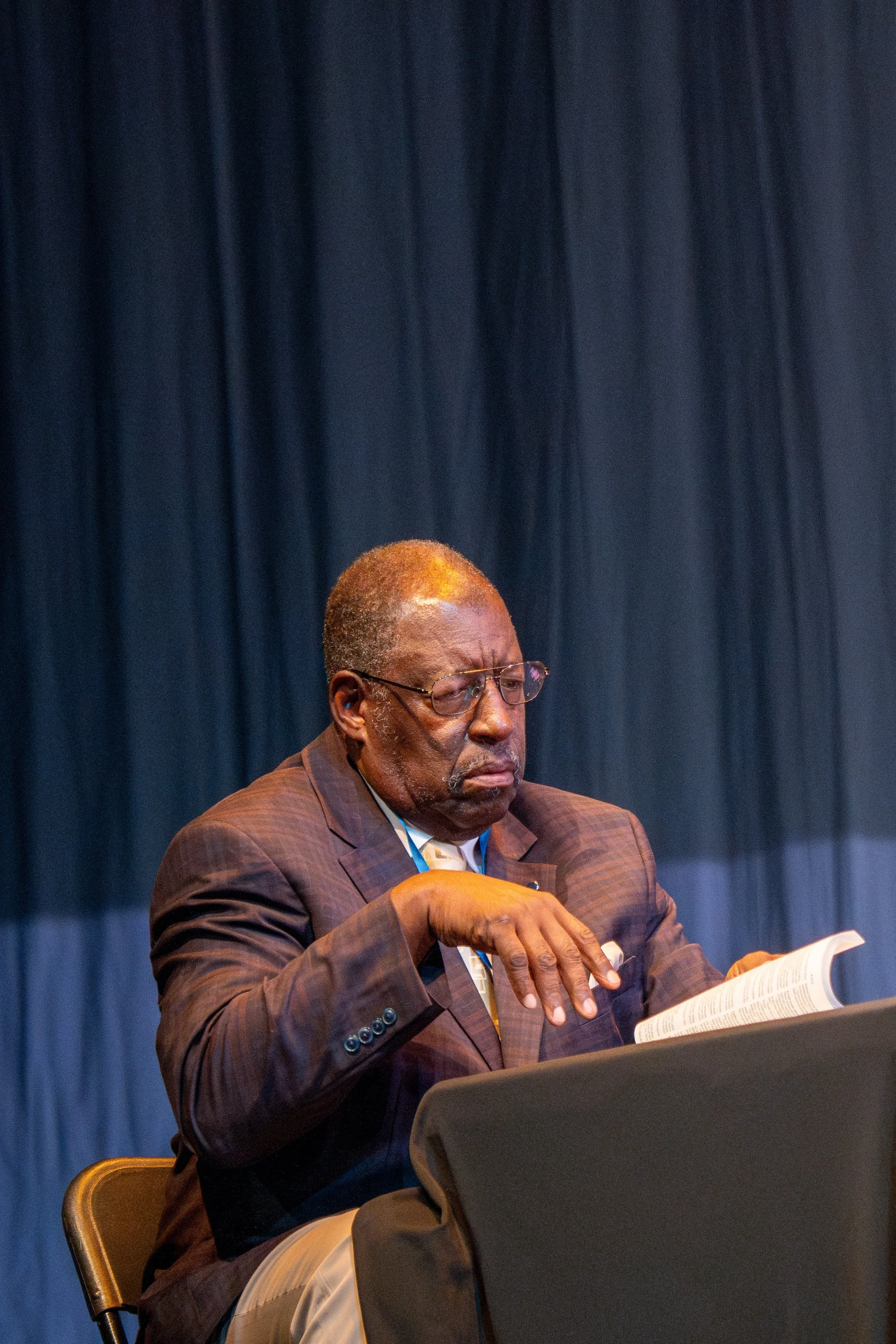 An elderly man with gray hair and glasses, wearing a brown checked blazer and cream pants, sitting at a table reading a book with a serious expression against a backdrop of a dark curtain.