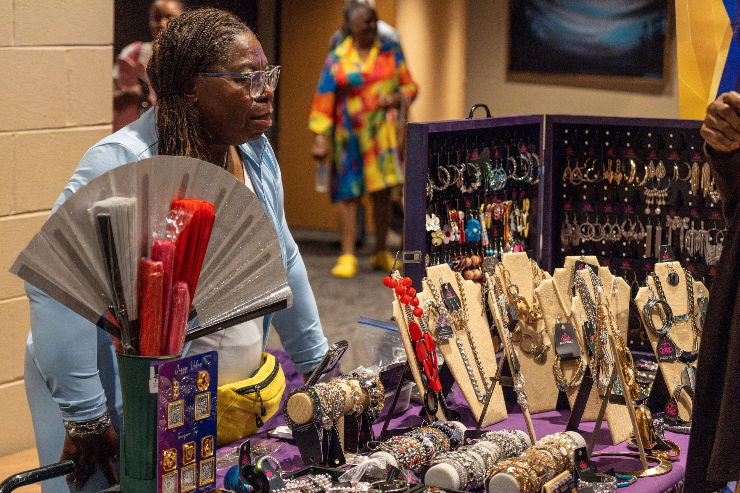 A woman with glasses browsing jewelry at a market stall with earrings, bracelets, and necklaces on display.
