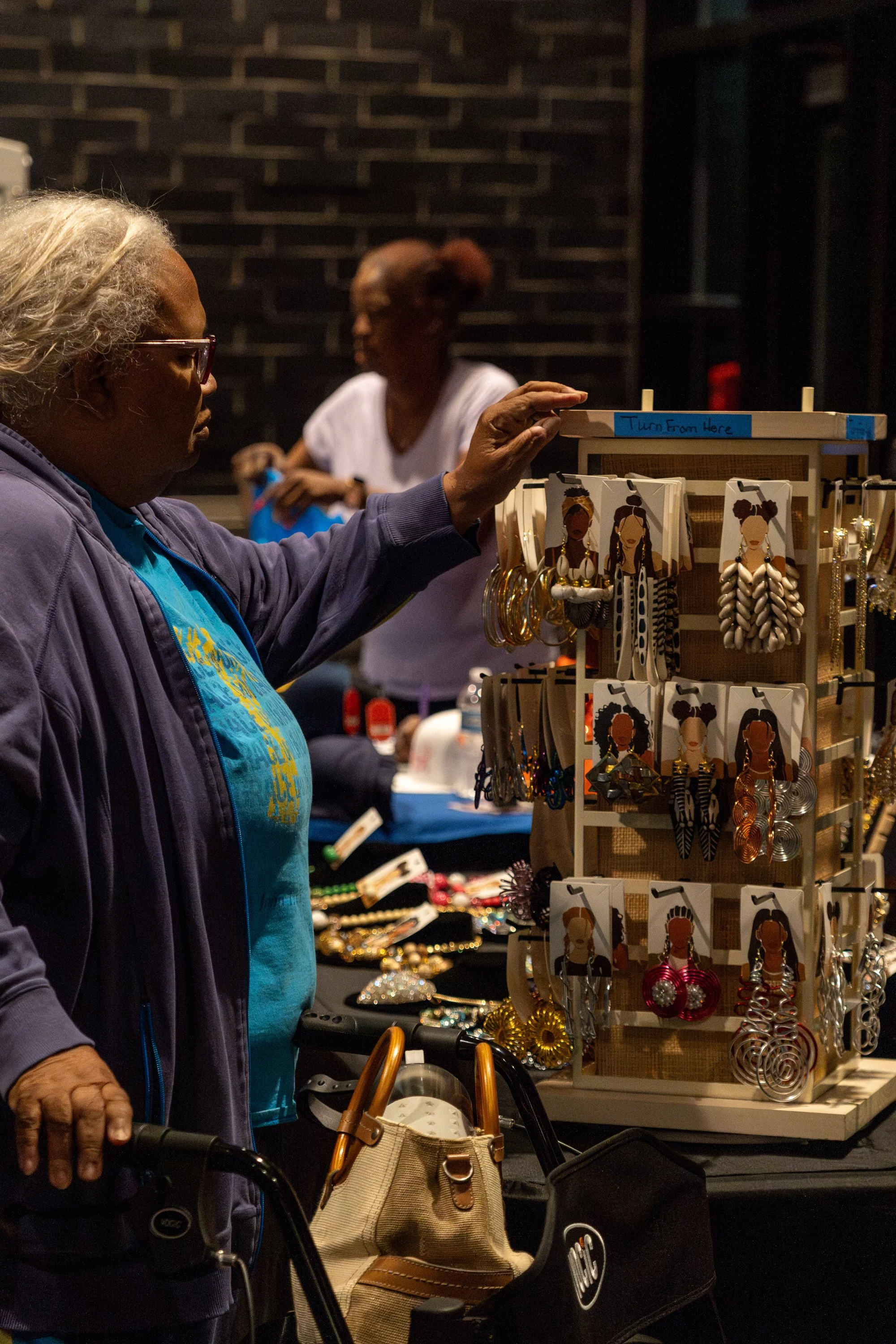 A woman with gray hair and glasses shopping for jewelry at an outdoor market stall at night. The stall displays earrings and necklaces featuring artwork of women with different hairstyles.