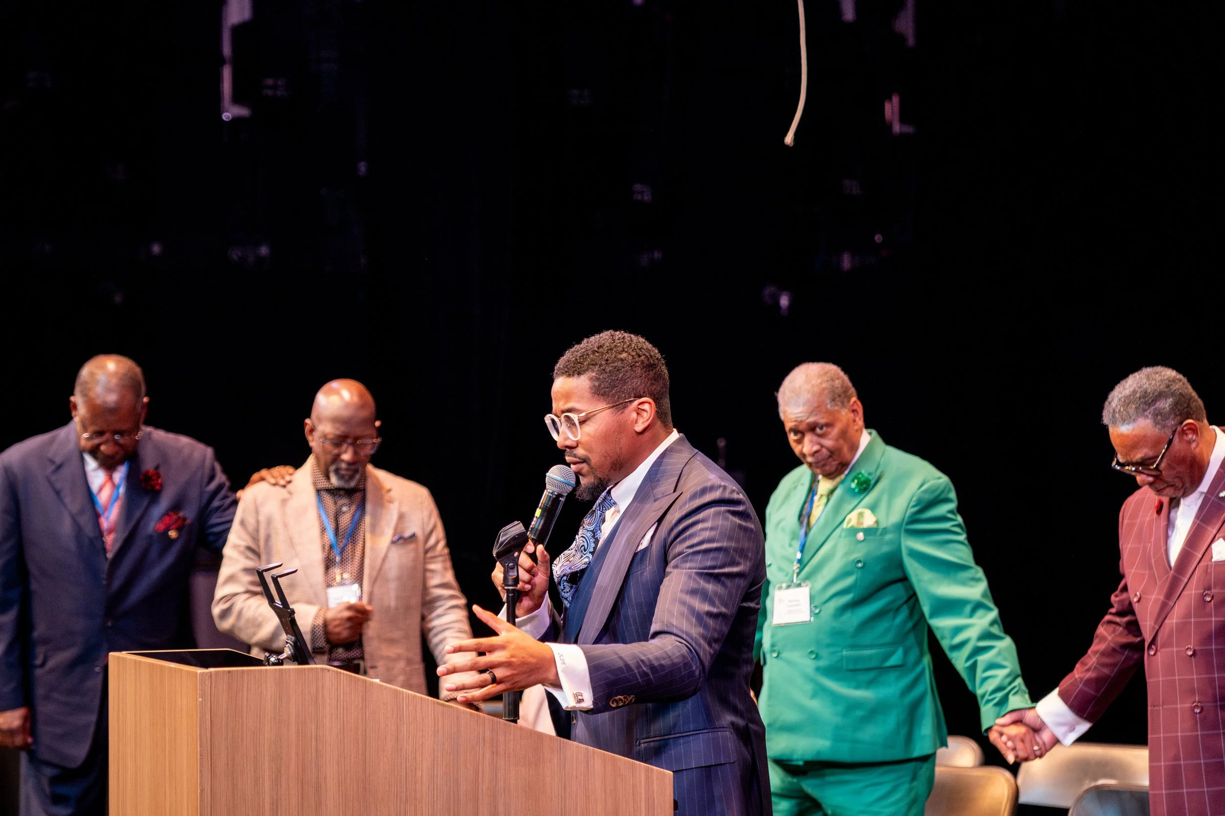 A group of men standing in a line on stage, with one man in the center speaking into a microphone, dressed in formal suits and colorful ties, at a formal event or conference.