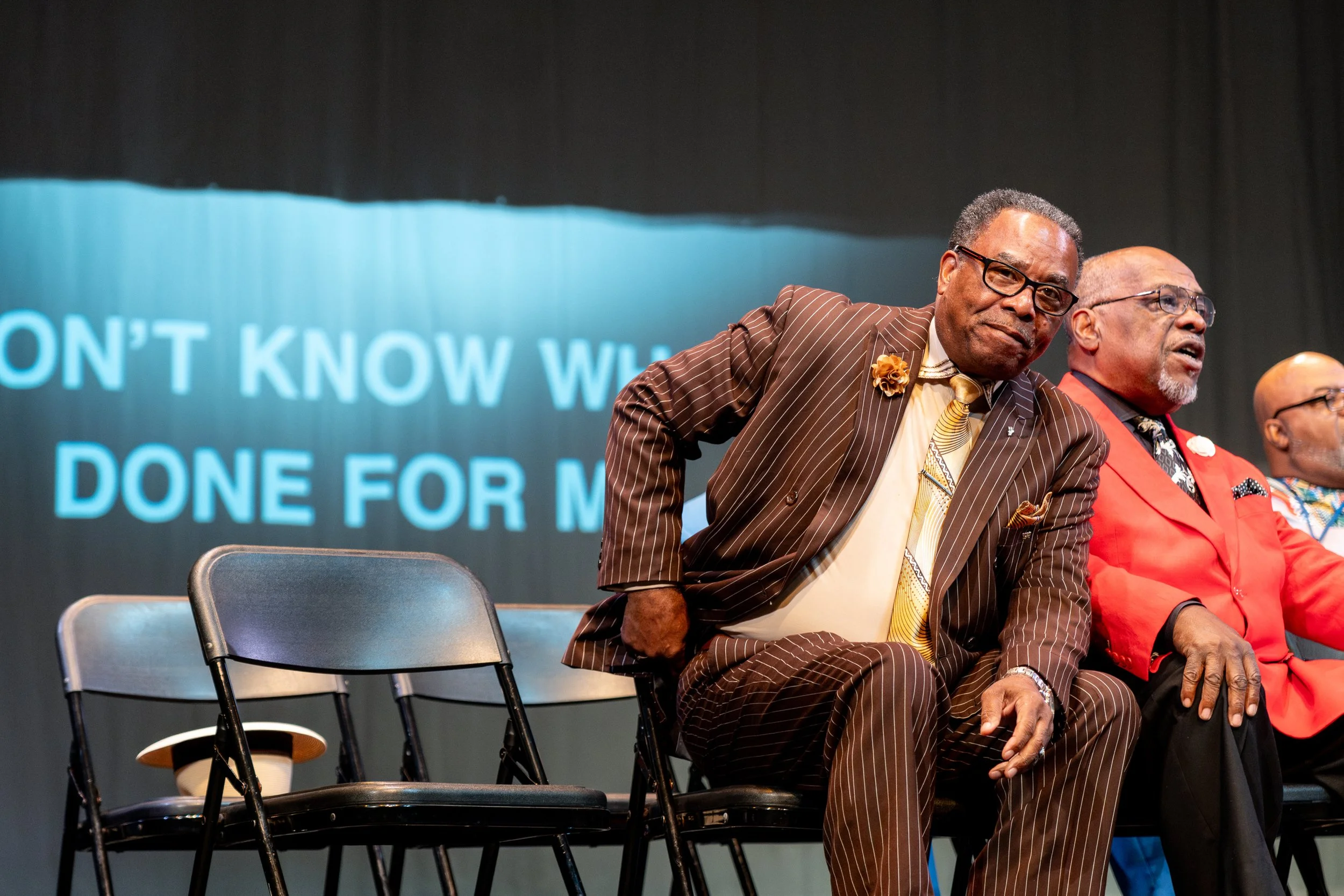 Three men sitting on chairs on a stage. The man in the foreground is wearing a brown striped suit with a flower pin, glasses, and a yellow tie. The man in the background is dressed in a red blazer with a colorful shirt. The third man, partly visible,