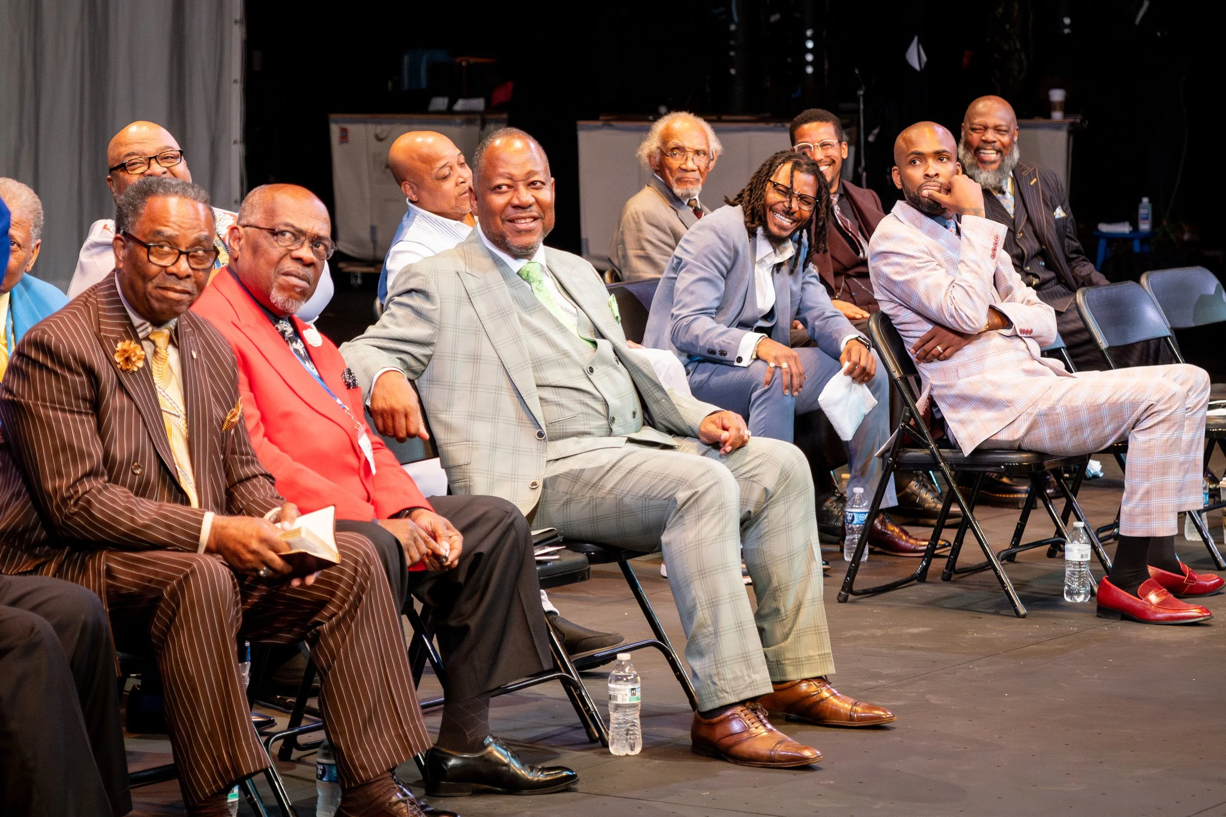 A group of diverse men, dressed in suits, sitting on chairs in a conference or event setting, some smiling and engaging, with water bottles on the floor.