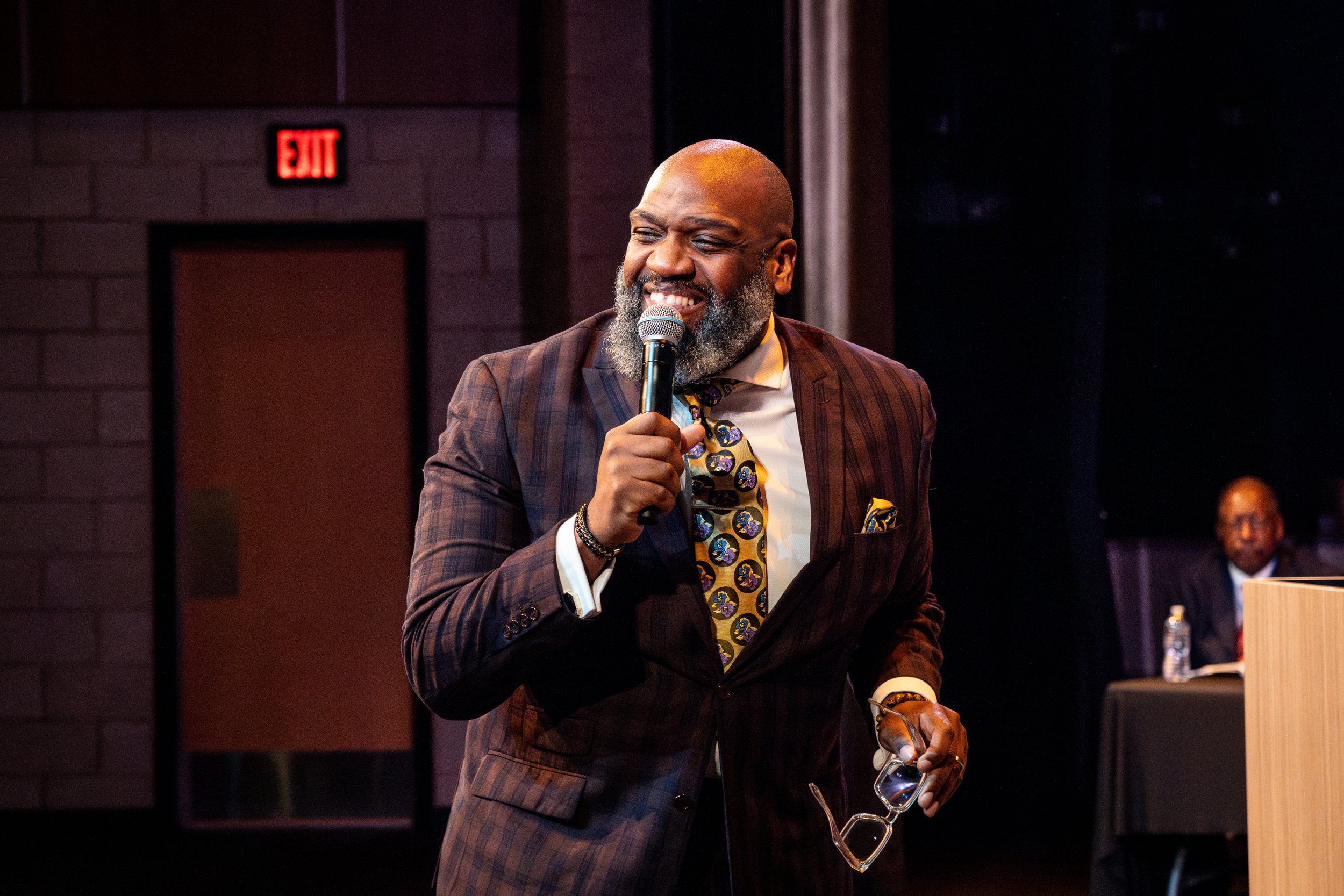 A man with a beard and bald head, smiling, wearing a brown plaid suit, holding a microphone and glasses, standing in front of a dark background during a speaking event.