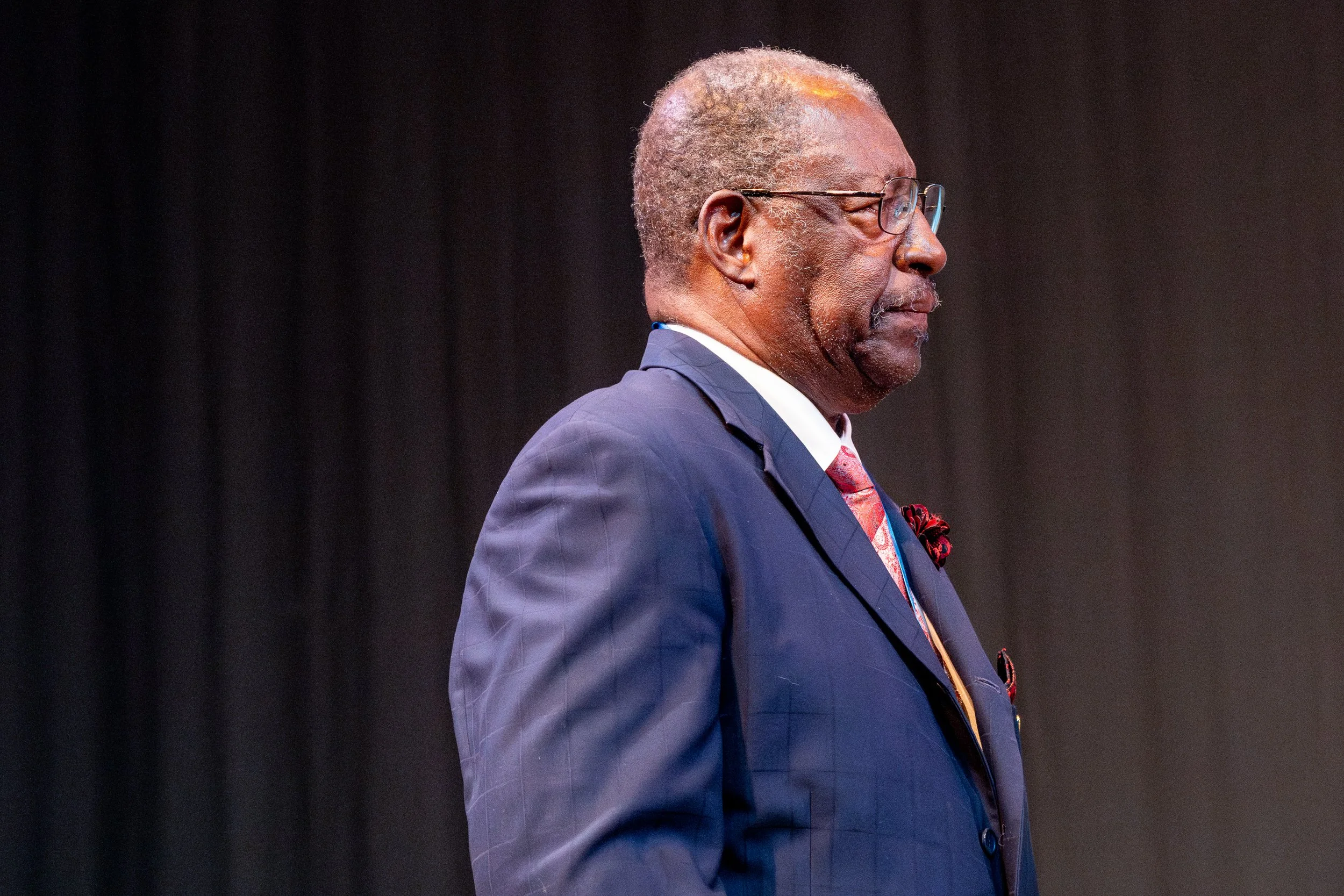 An elderly man wearing glasses, a dark blue checkered suit, and a red tie standing against a dark background.