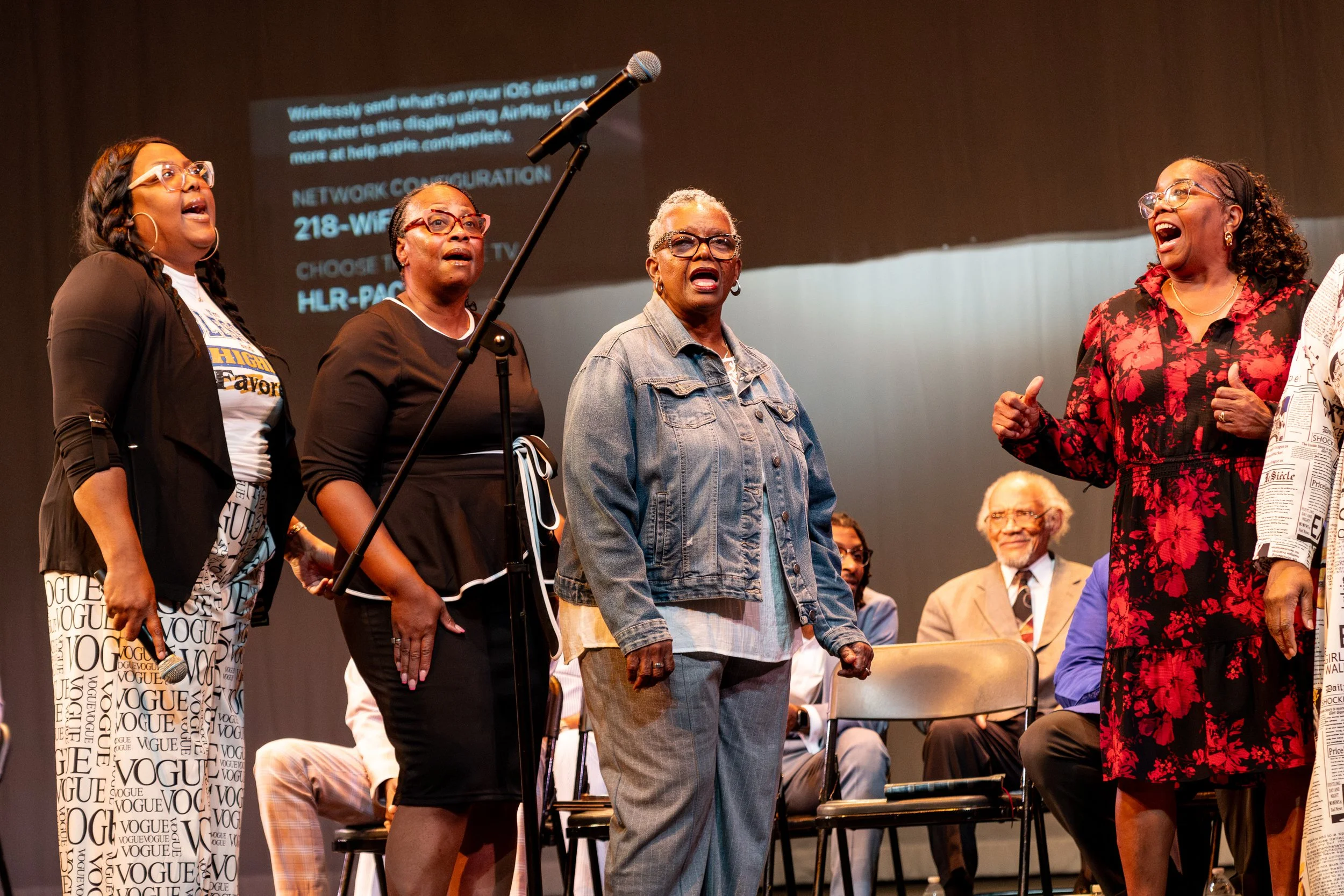 A group of diverse women standing on stage and singing or speaking into a microphone in front of an audience. The women are dressed in various styles, including casual and patterned clothing.