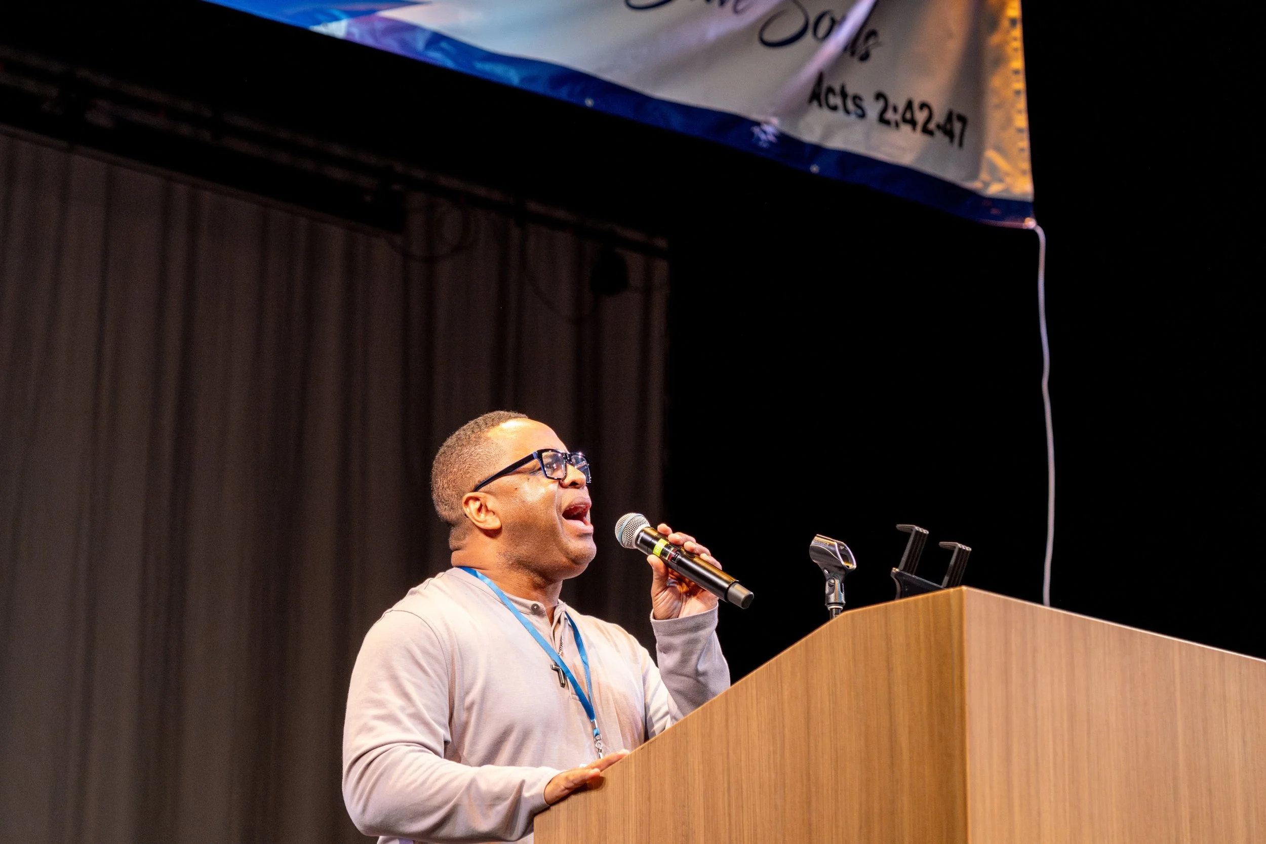 A man with glasses speaking or singing into a microphone at a podium during an indoor event.