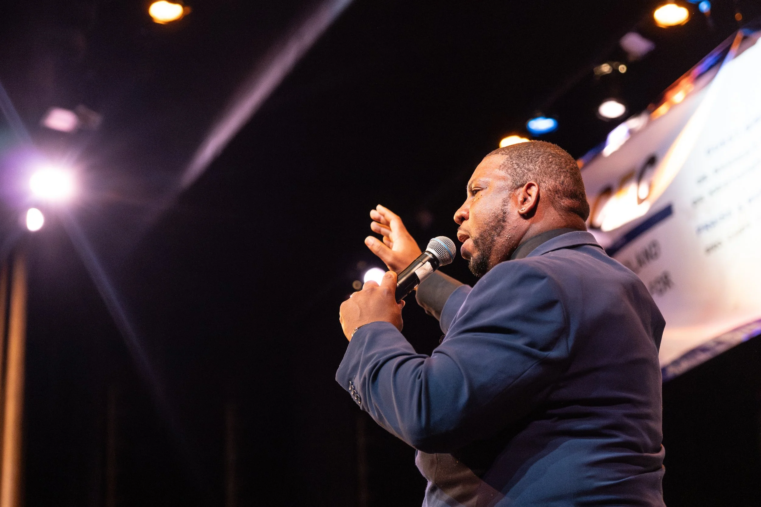 A man in a dark jacket speaking into a microphone, with a presentation or screen in the background. The setting appears to be an indoor event or conference with stage lighting.