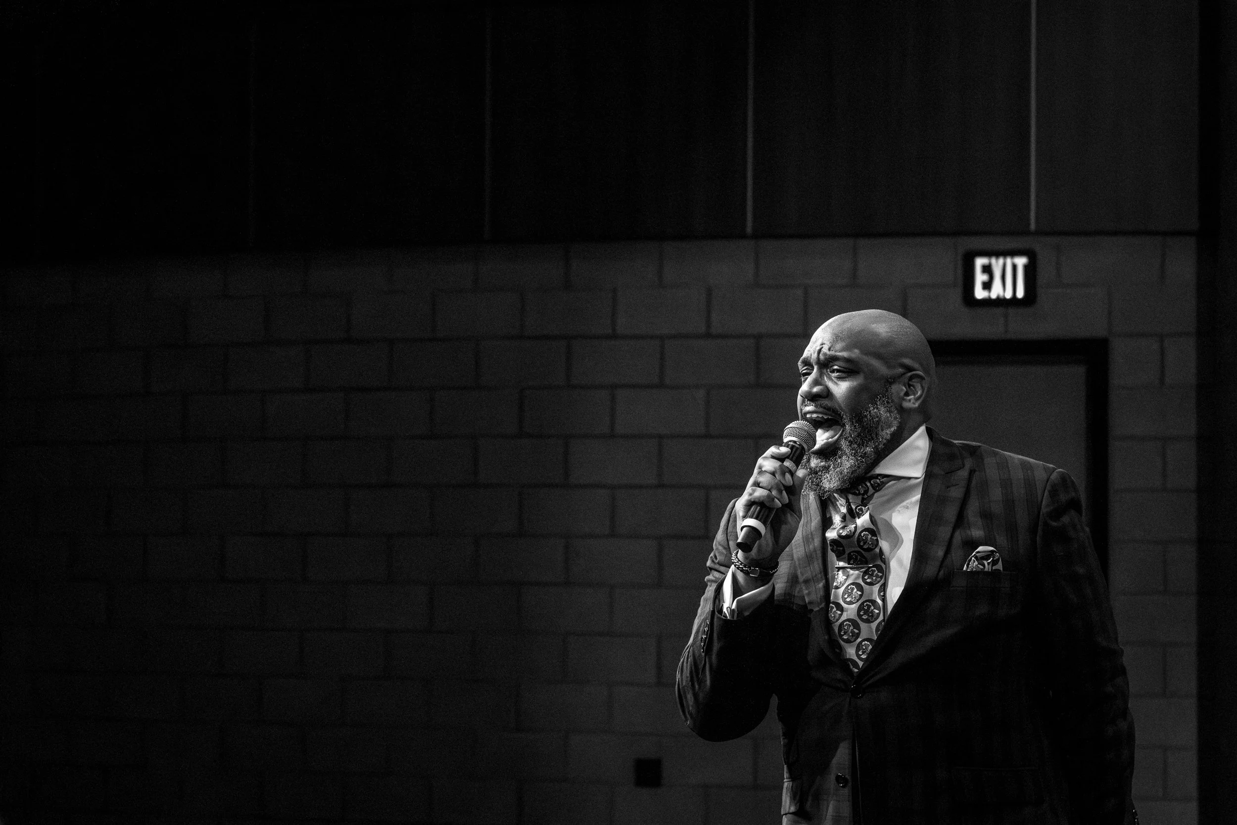 A Black man singing into a microphone on stage, dressed in a checked suit, polka dot tie, and patterned shirt, against a dark brick wall background with an exit sign.