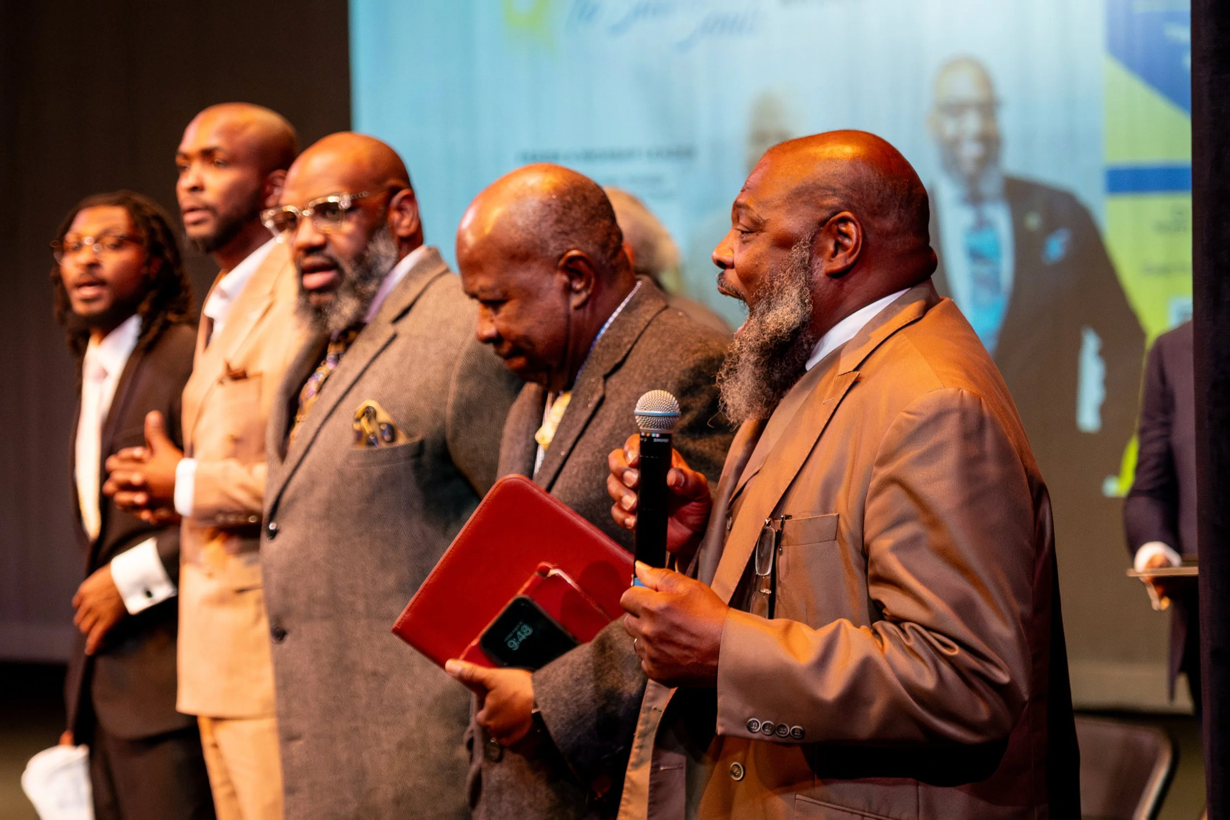 Six men in suits standing in a row during a conference or panel discussion. One man is holding a microphone and a red folder. A large screen with a blurry image is in the background.