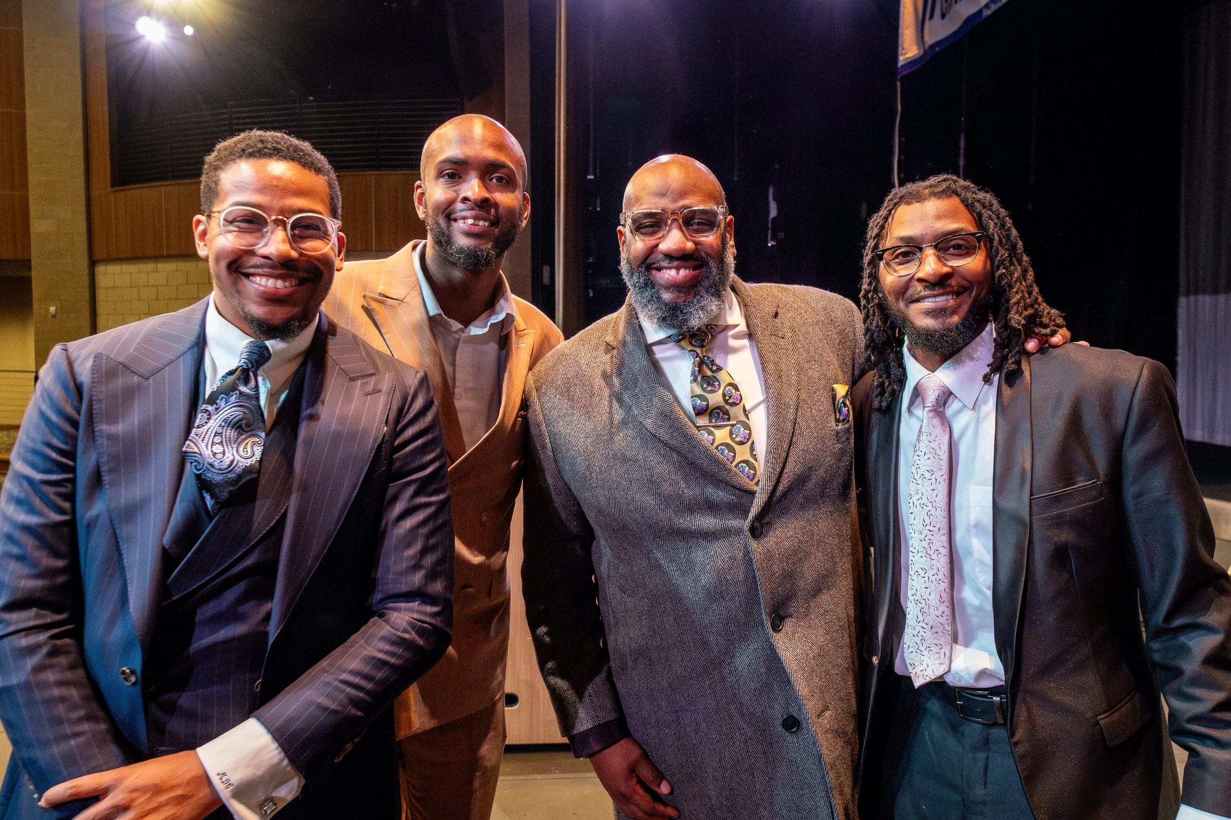 Four men in suits smiling at a camera, standing together in an auditorium or theater.