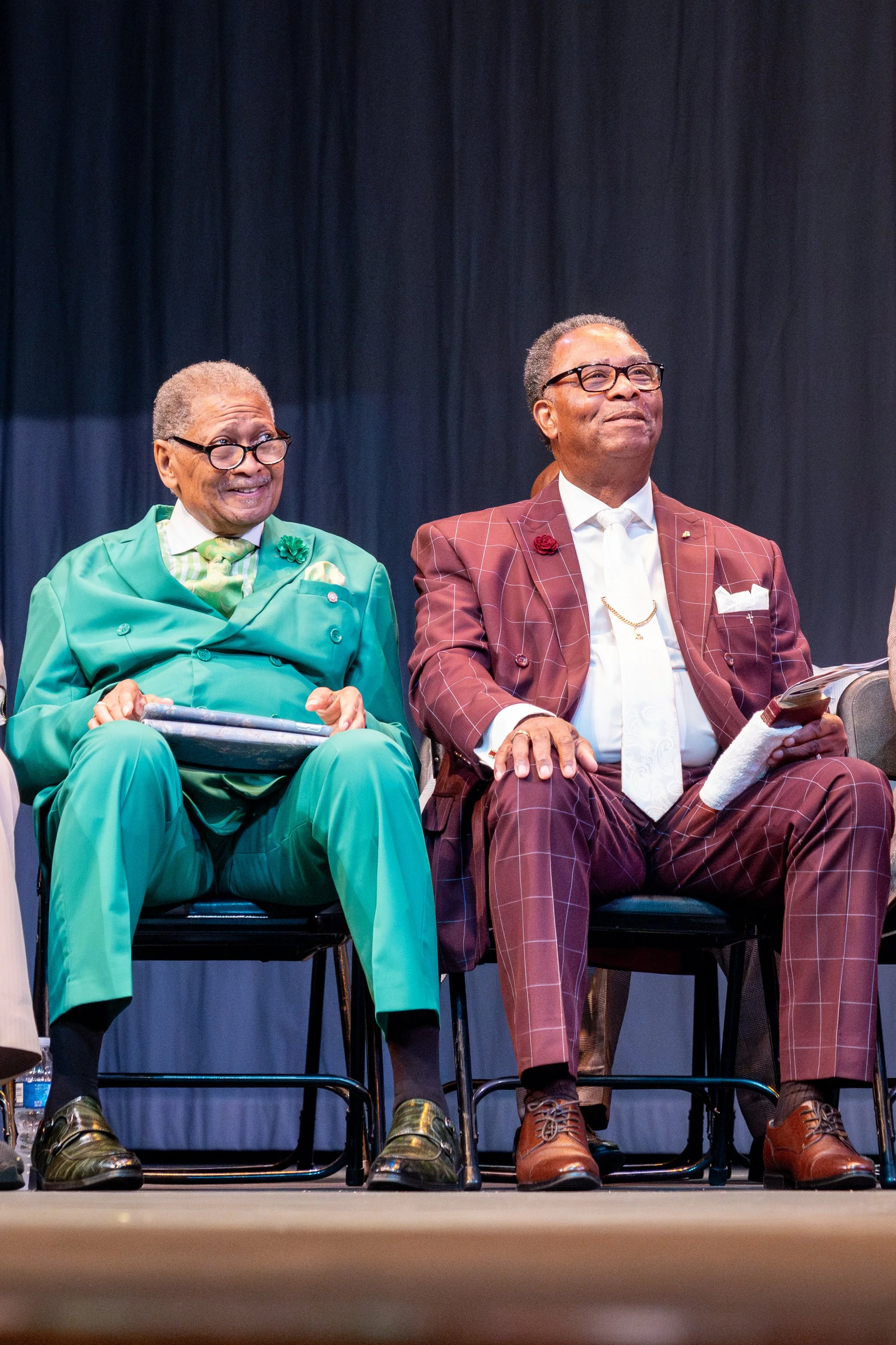 Two older men sitting on stage in chairs. The man on the left is wearing a bright green suit with a patterned shirt and glasses. The man on the right is dressed in a maroon checkered suit with a white shirt, glasses, and is holding papers. Both appea
