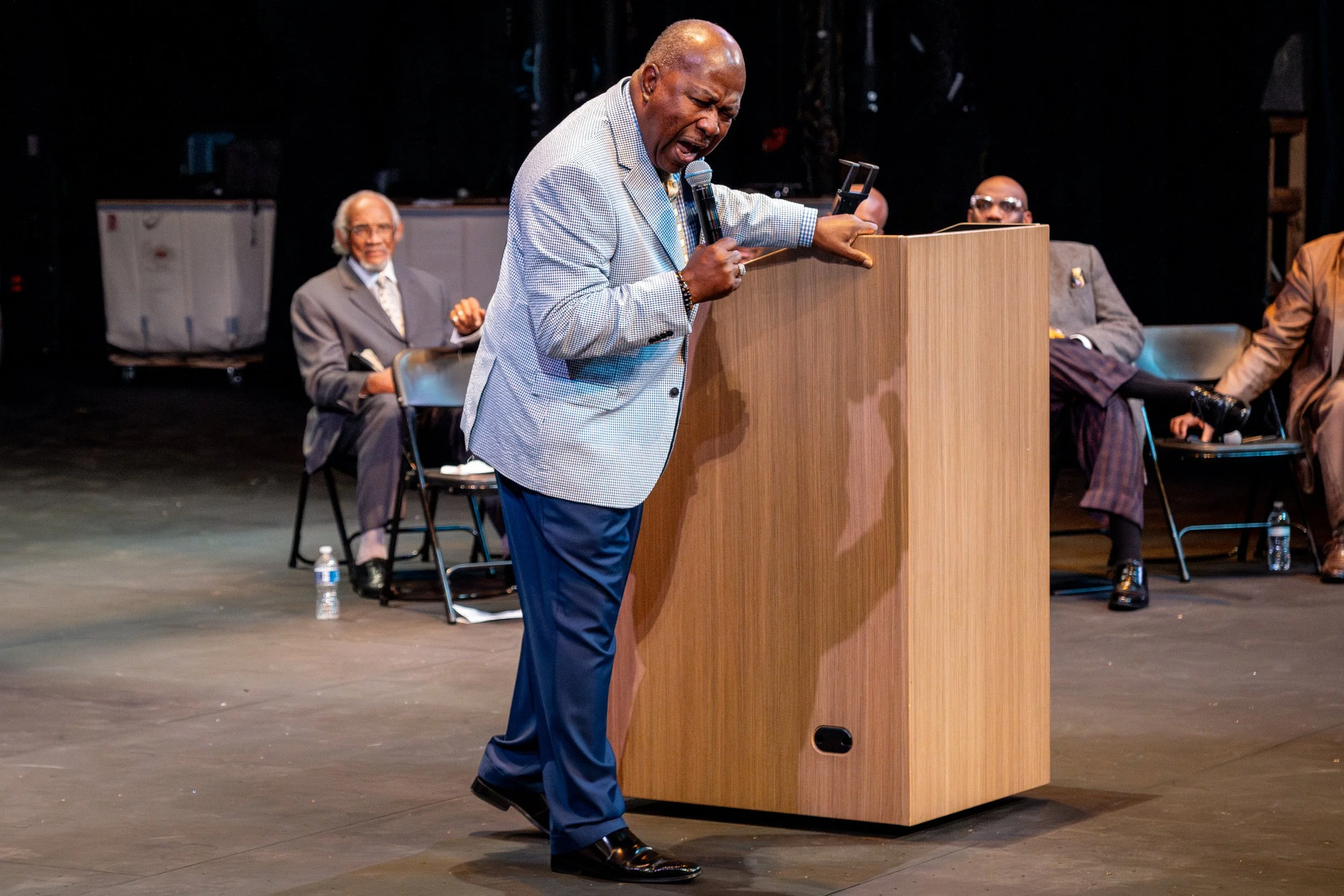 An elderly man in a checkered blazer passionately speaking at a podium on stage, with several seated men in suits behind him.