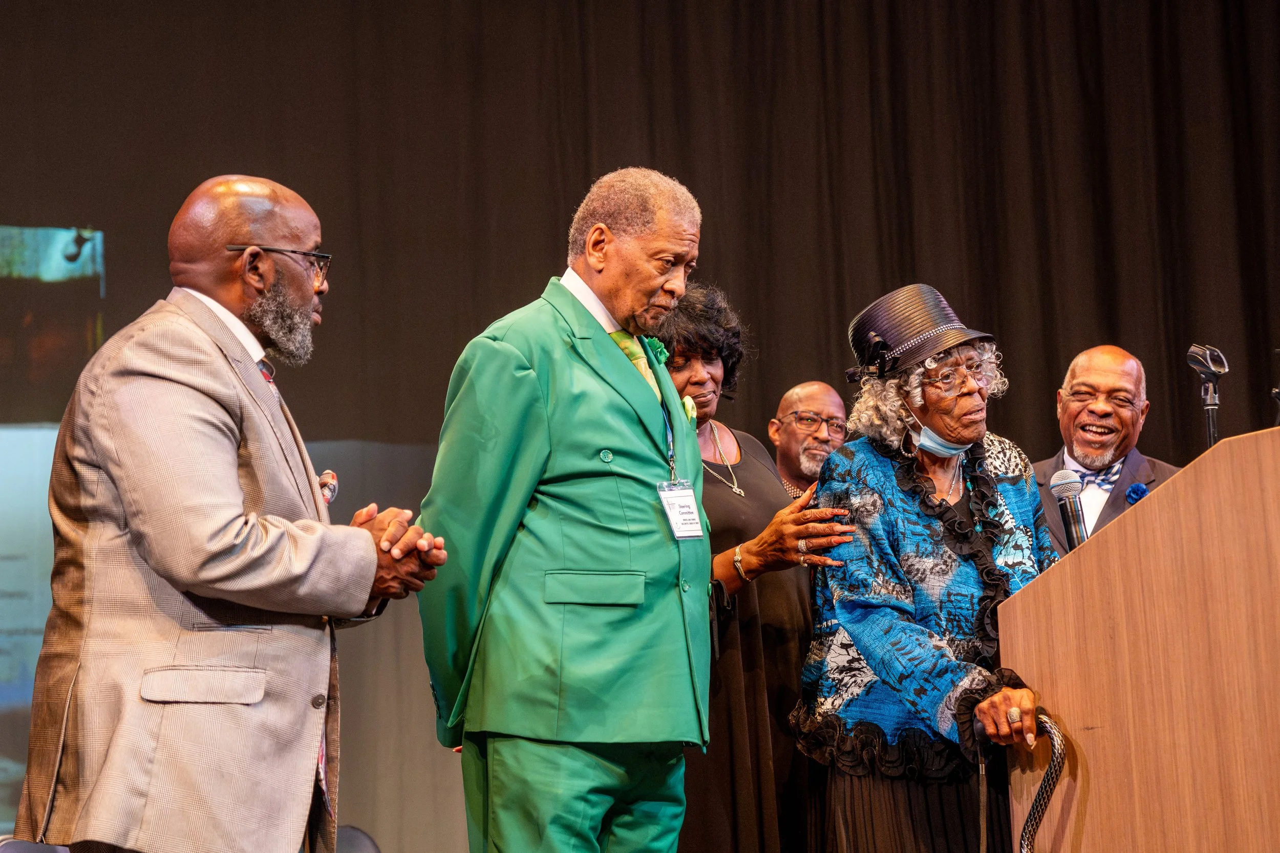 A group of six diverse older adults standing at a podium during a formal event or ceremony, with a dark curtain background.