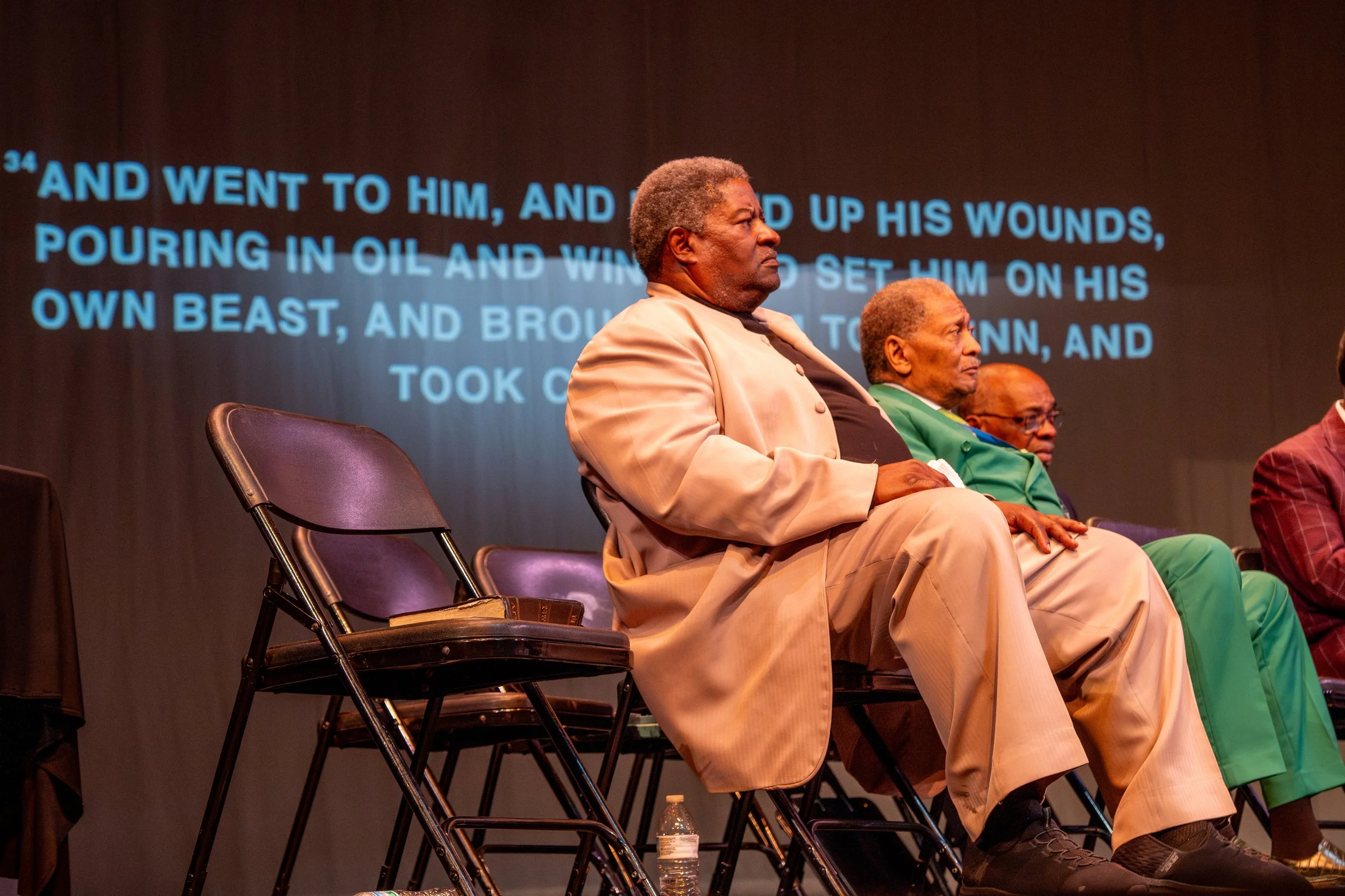 Three men sit on chairs during a presentation or panel discussion, with a large screen behind them displaying text. The first man, with gray hair, wears a beige suit and black shirt. The second man, with gray hair, wears a green blazer with a yellow 