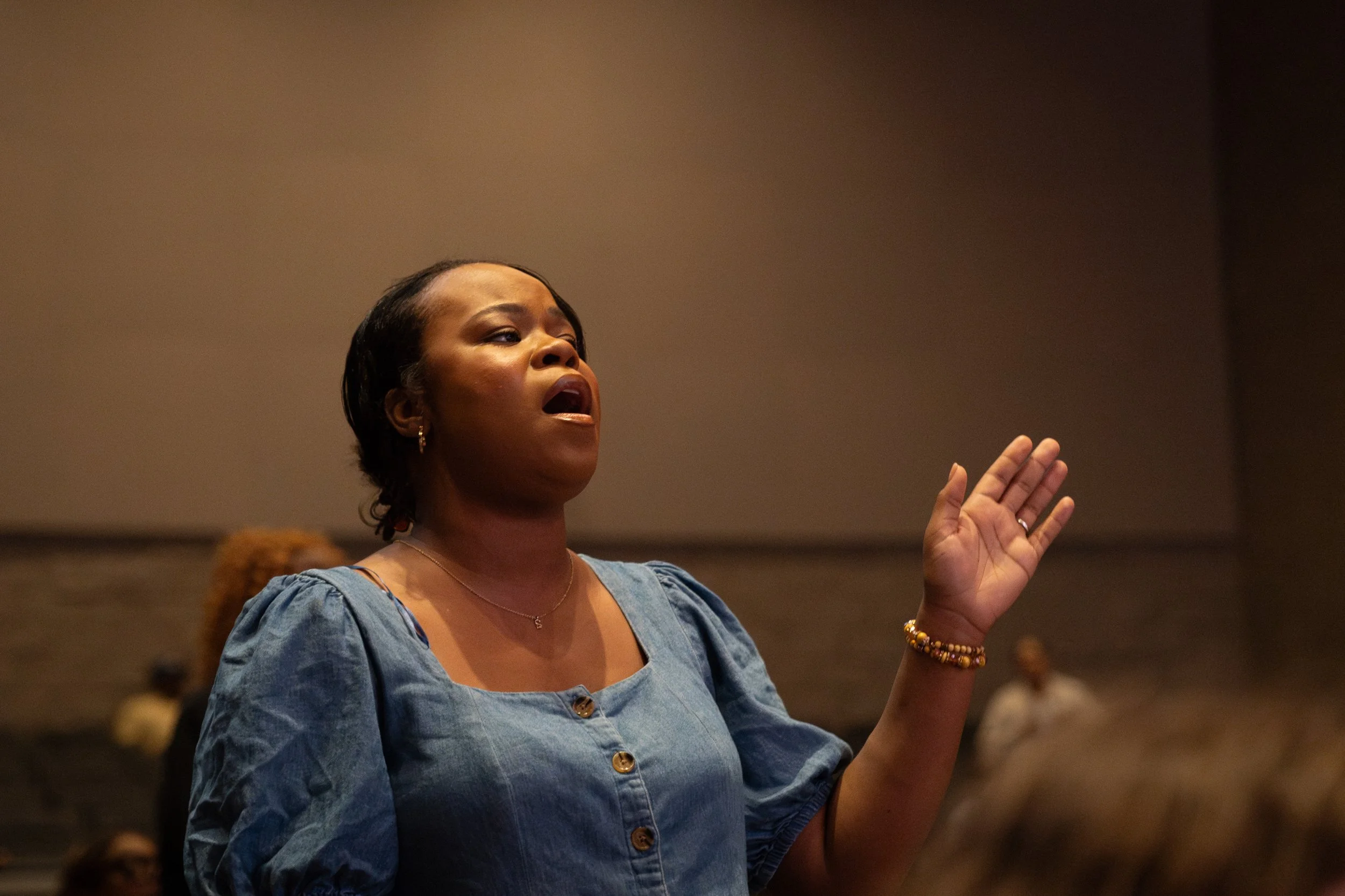A woman with short dark hair, wearing a blue blouse, appears to be singing or speaking with her eyes closed and her hand raised, in a dimly lit indoor setting.