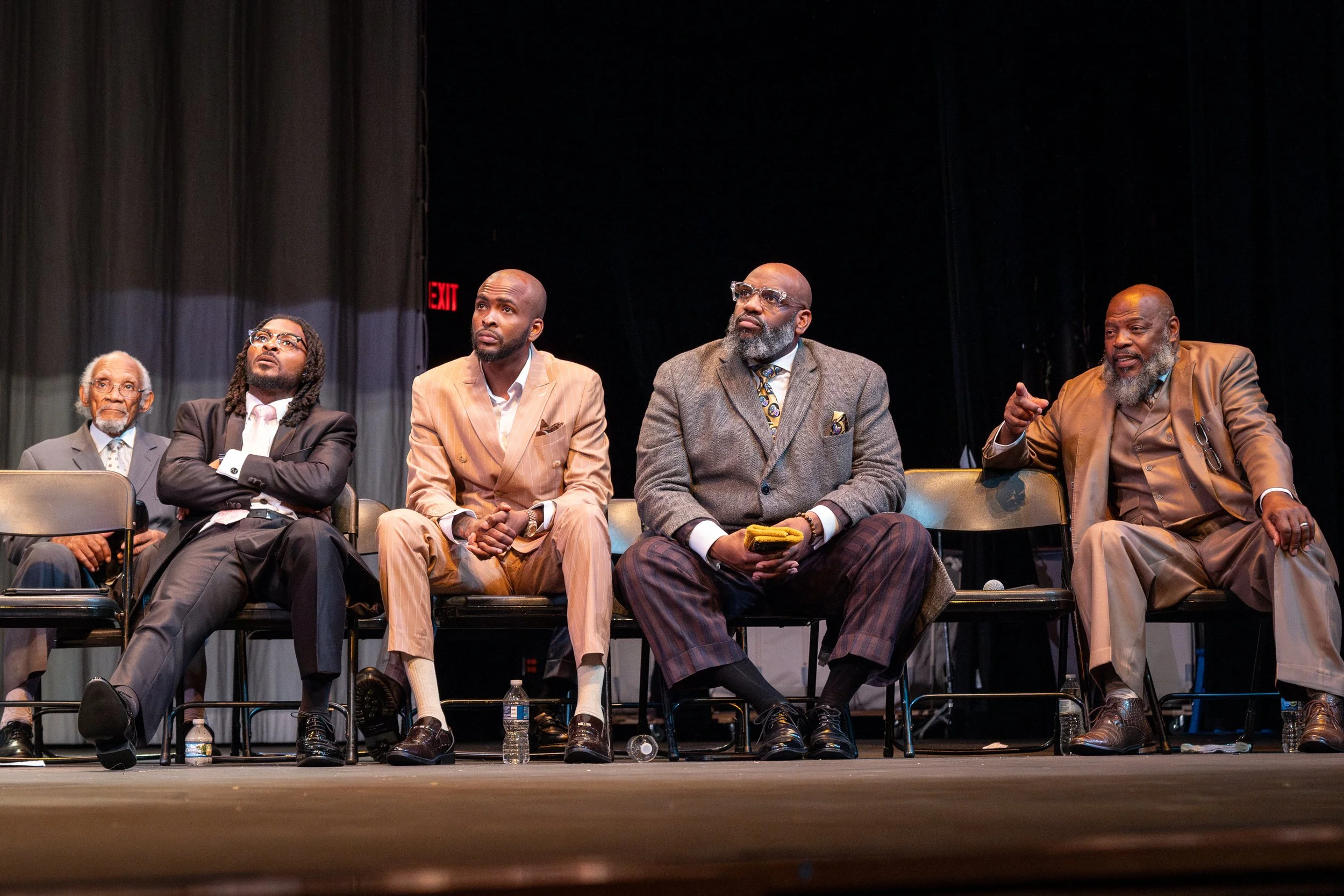 A group of six men sitting on a stage with black curtains and a red exit sign in the background, engaged in a panel discussion or presentation.