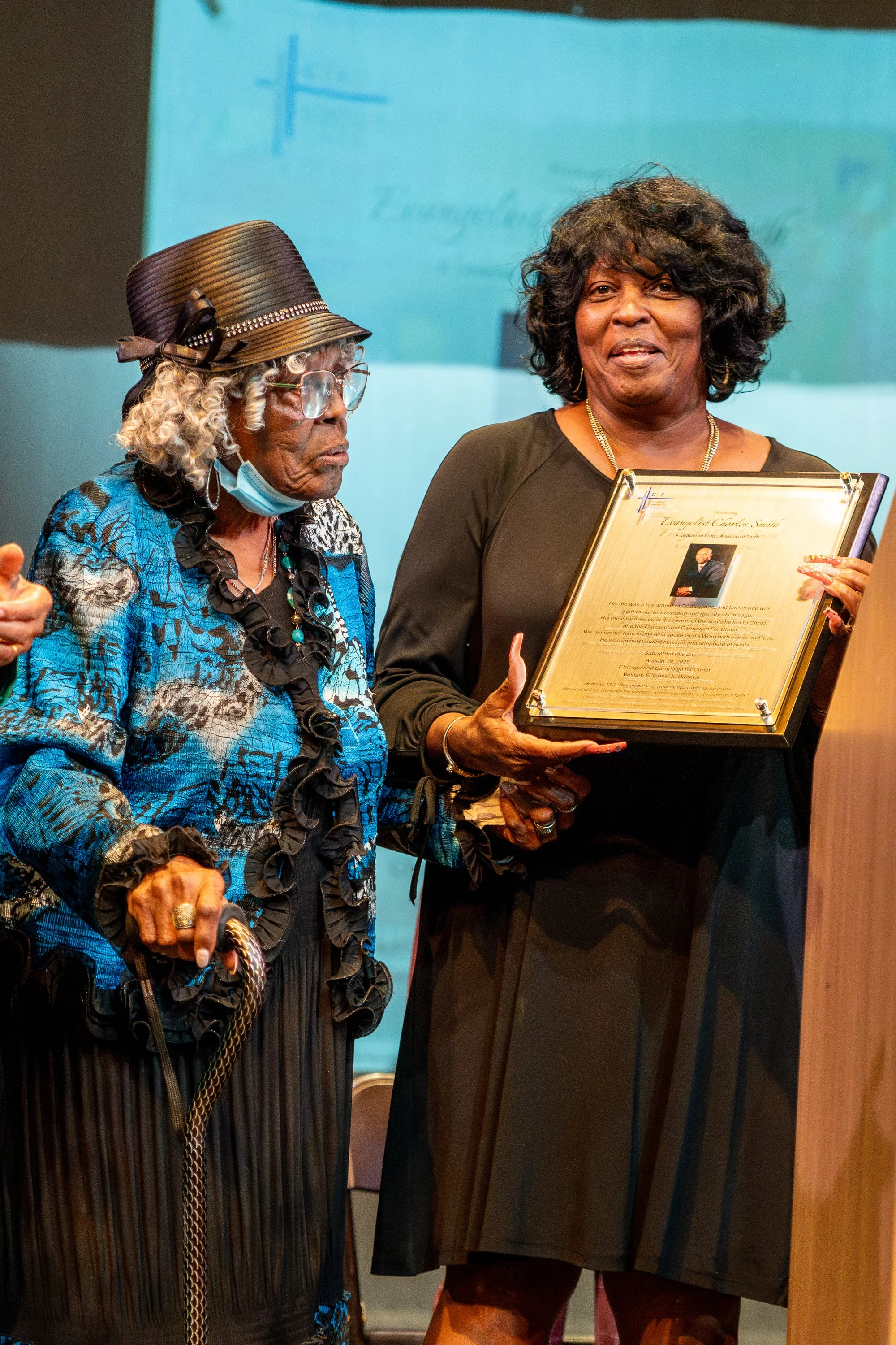 Two women, one older with gray curly hair wearing glasses, a blue floral dress, and a large hat, the other younger with black curly hair wearing a dark dress, holding a framed certificate, standing together at an indoor event.