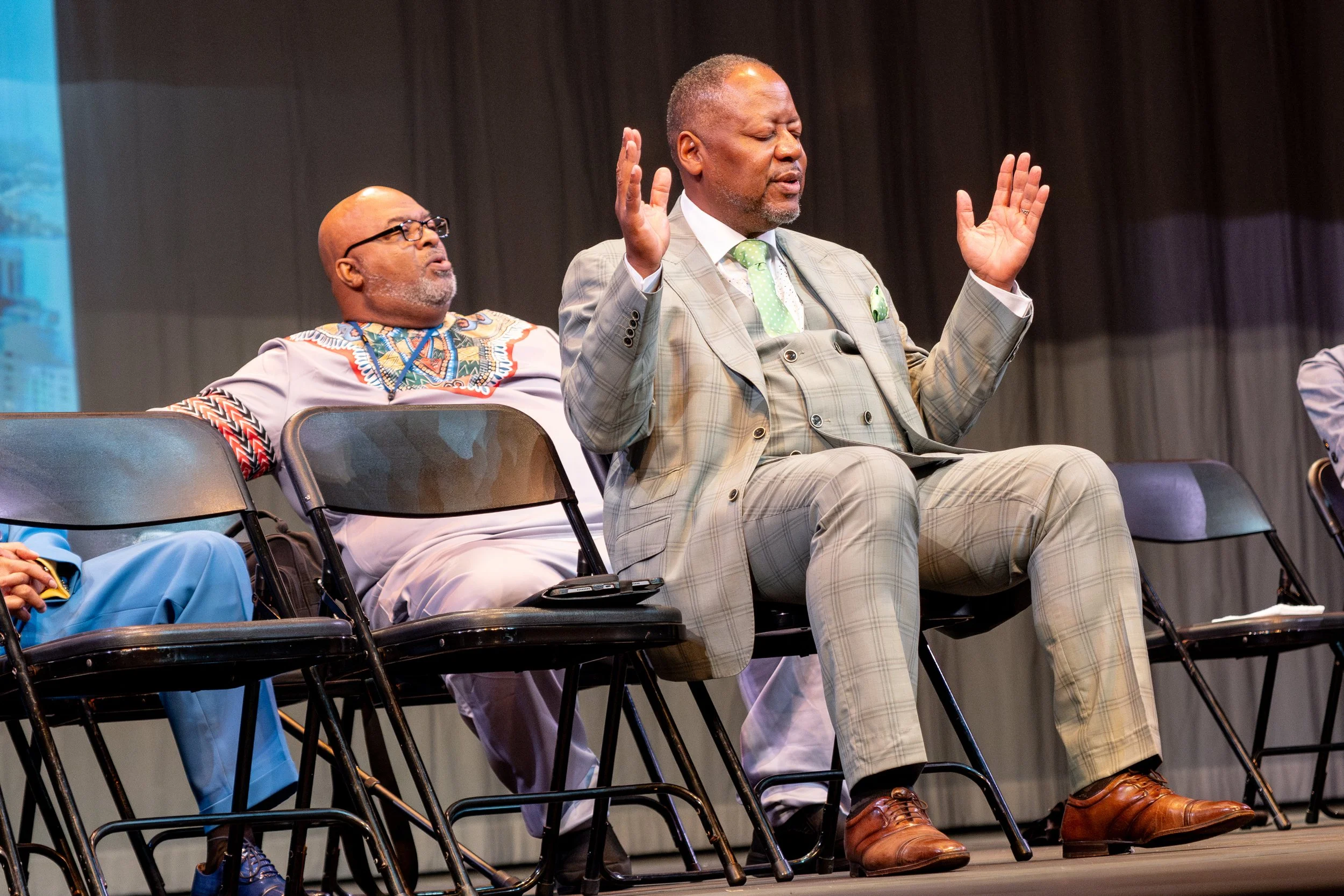 Three men sitting on stage during a formal event. The man in the middle is praying with his eyes closed and hands raised. The man on the left is wearing glasses and traditional African attire, looking up. The man on the right is wearing a light-color