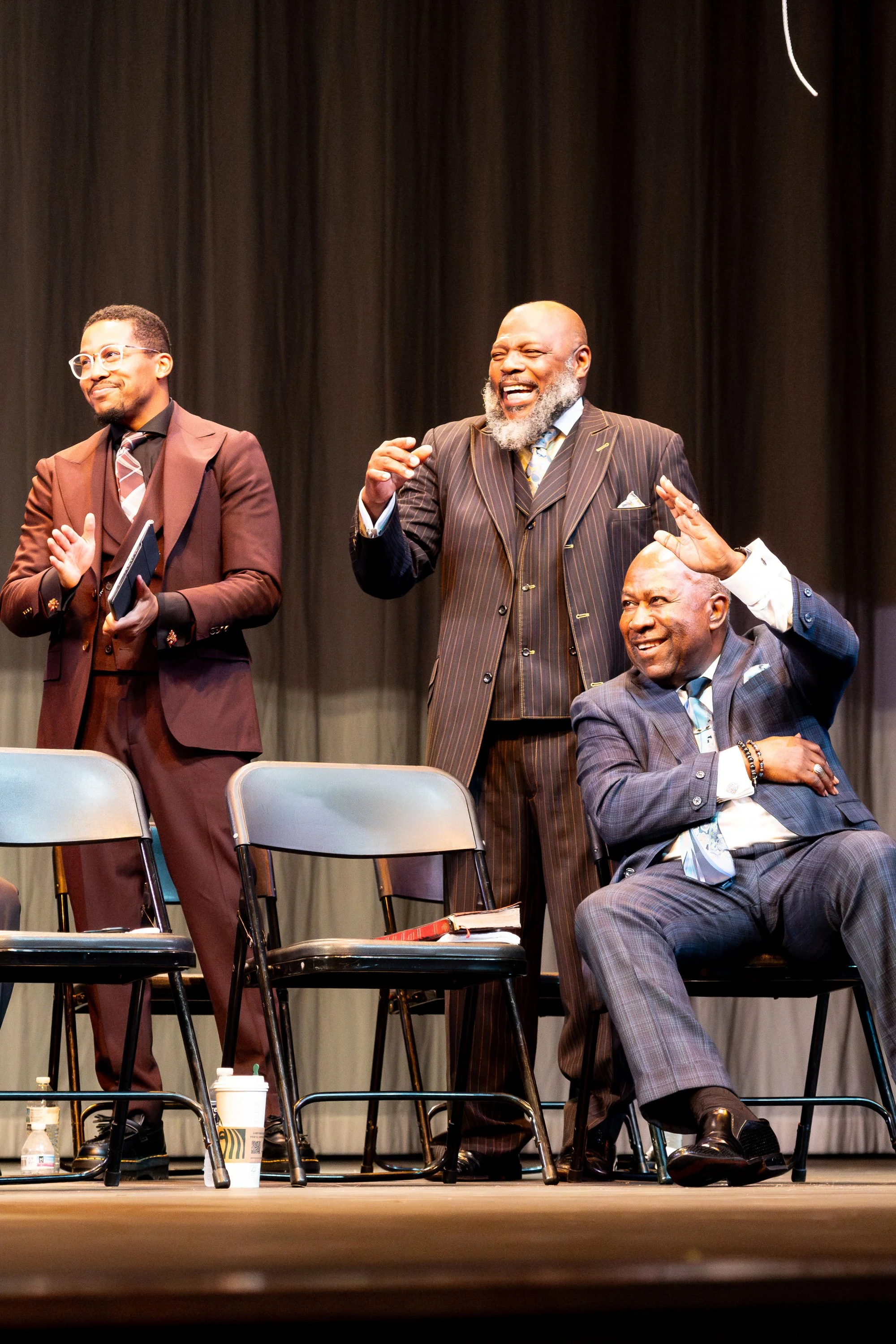 Three men in suits on stage, two standing and one seated, laughing and engaging with each other. The stage has chairs and a table with drinks and supplies, with a dark curtain backdrop.