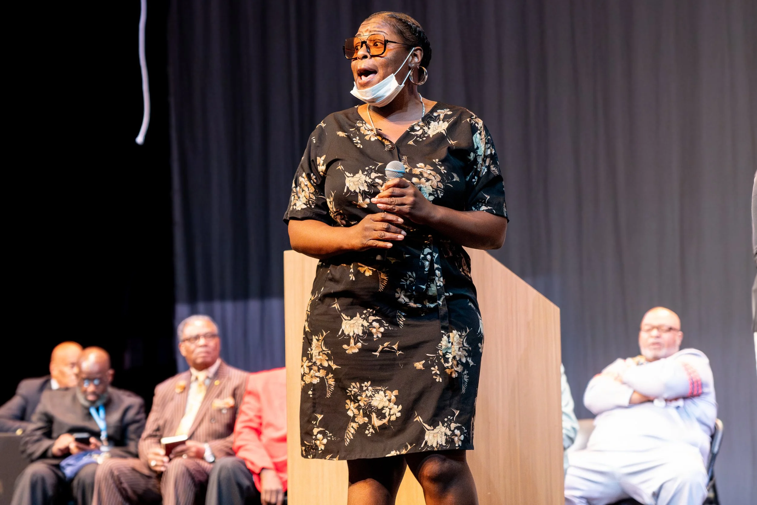 A woman in glasses and a floral dress speaking into a microphone on stage with a black curtain background. Several seated men are visible in the background.