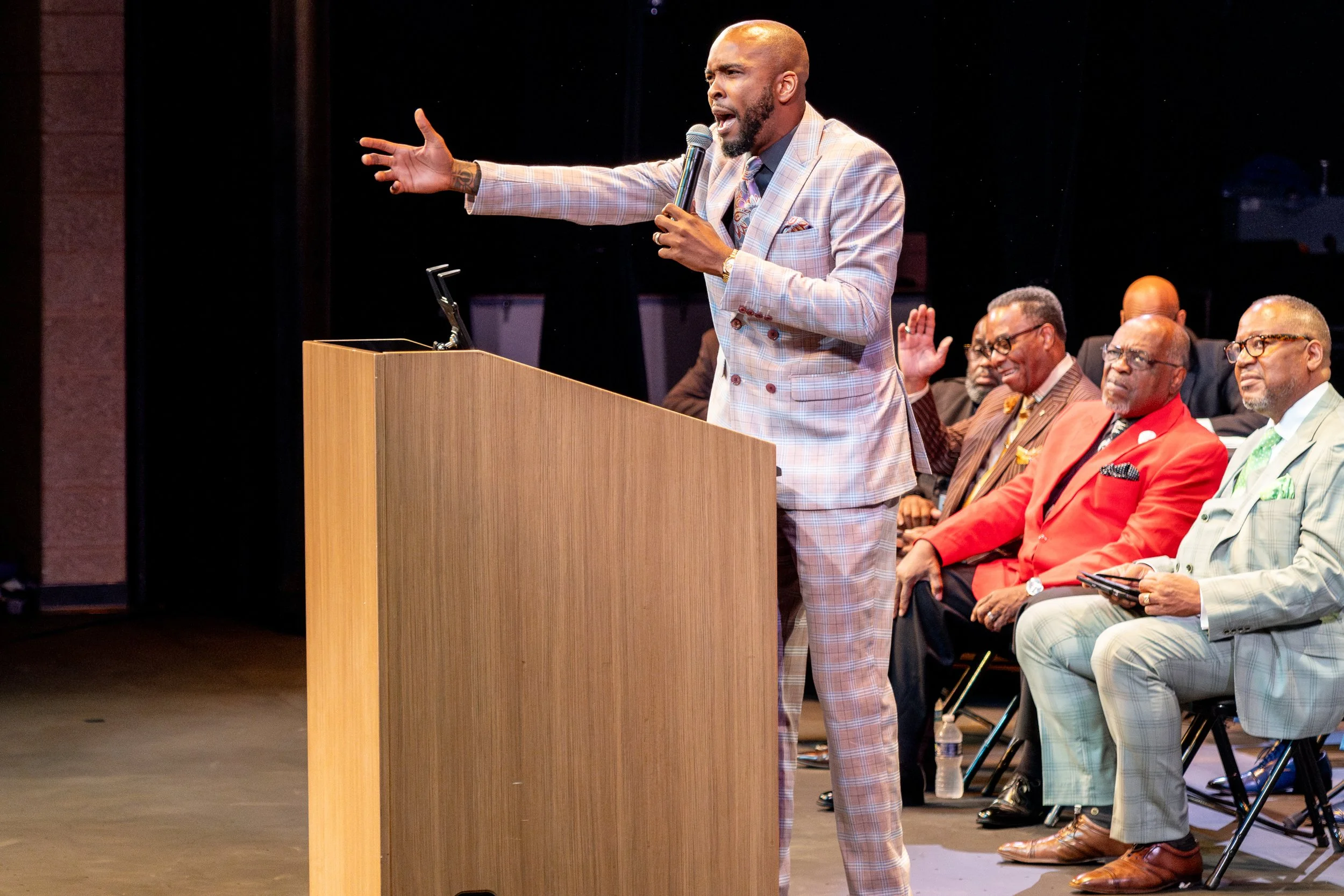 A man in a light-colored plaid suit speaking passionately at a podium with a microphone, while seated individuals watch, some with hands raised, in a darkened auditorium.