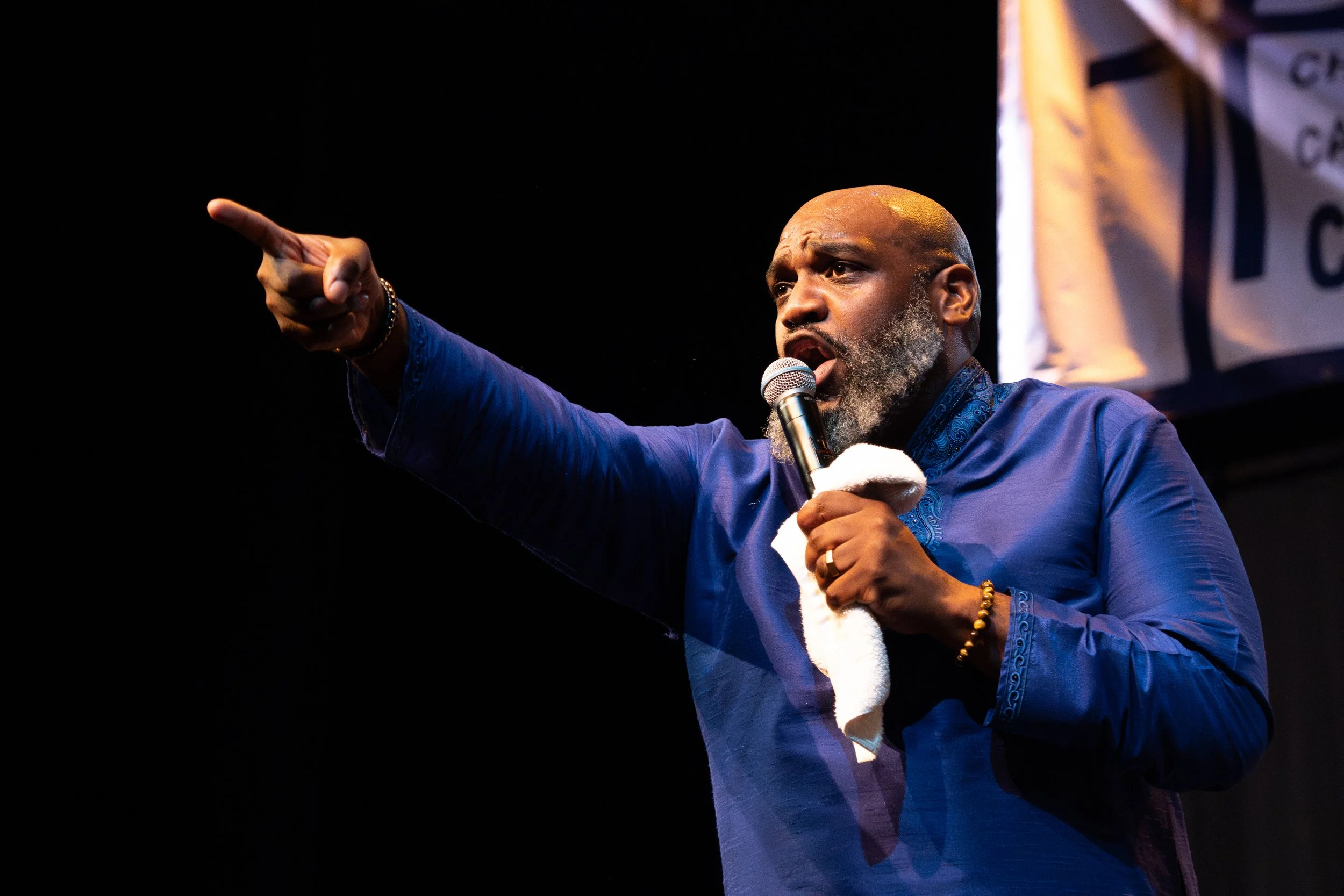 A man with a bald head and gray beard speaking into a microphone on a stage, pointing with his right hand, wearing a blue shirt, and holding a towel or cloth in his left hand.