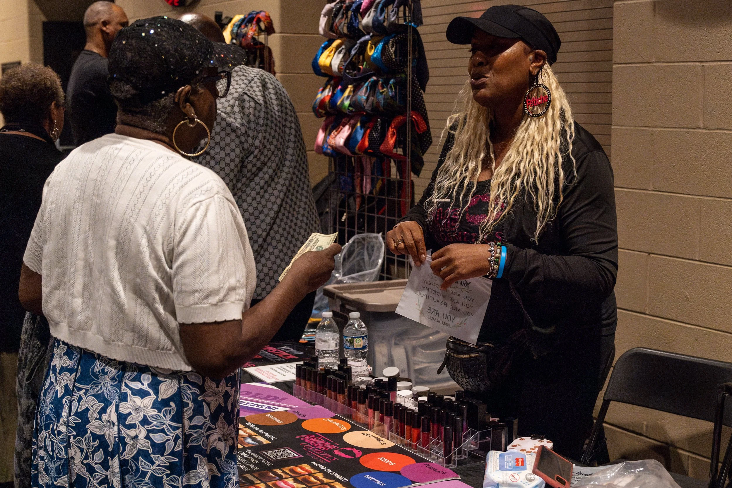 A woman with long blonde curly hair, wearing a black cap and large earrings with the word 'Revolution' in red, is talking to an older woman at a vendor booth. The booth has various makeup products, including lipsticks, and informational materials. Ot