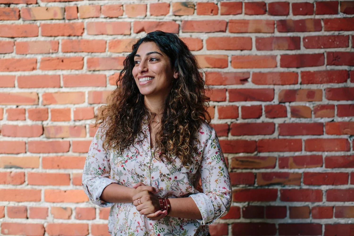 A woman with long, curly brown hair, smiling and looking to her left, stands in front of a red brick wall. She is wearing a light-colored, floral-patterned shirt with the sleeves rolled up, a bracelet, and a ring.