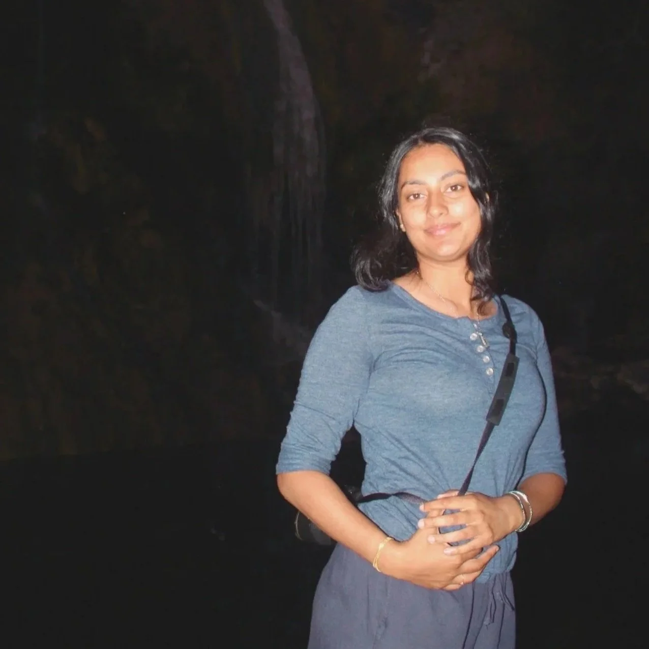 A woman with black hair wearing a blue long-sleeve top and gray pants, standing in front of a dark cave or waterfall environment during the night.