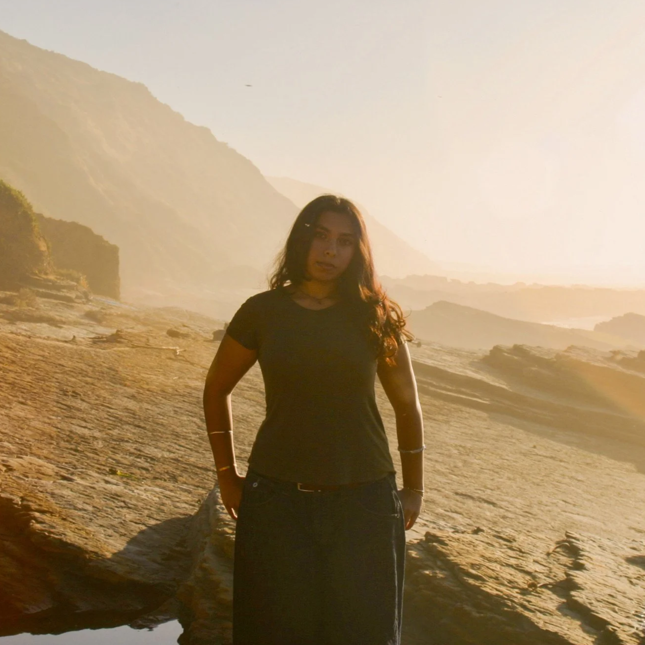 A woman standing on a rocky beach during sunset or sunrise, with mountainous terrain in the background and warm golden light illuminating the scene.