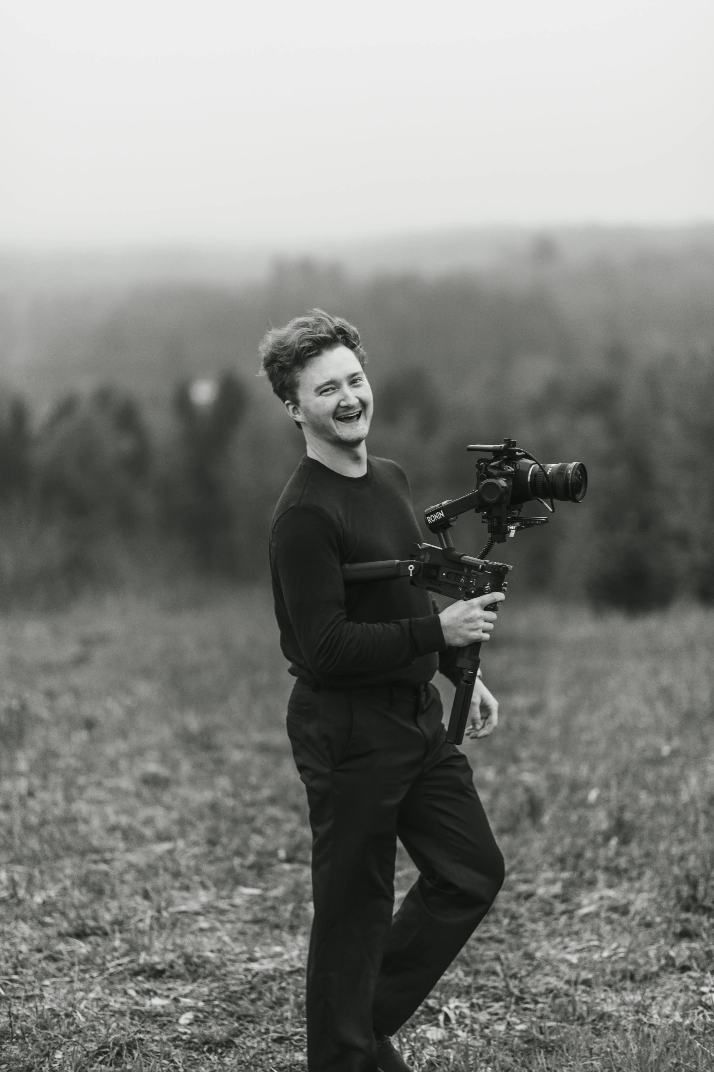 A young man with curly hair smiling and holding a camera on a stabilizer outdoors in a field, black and white photograph.