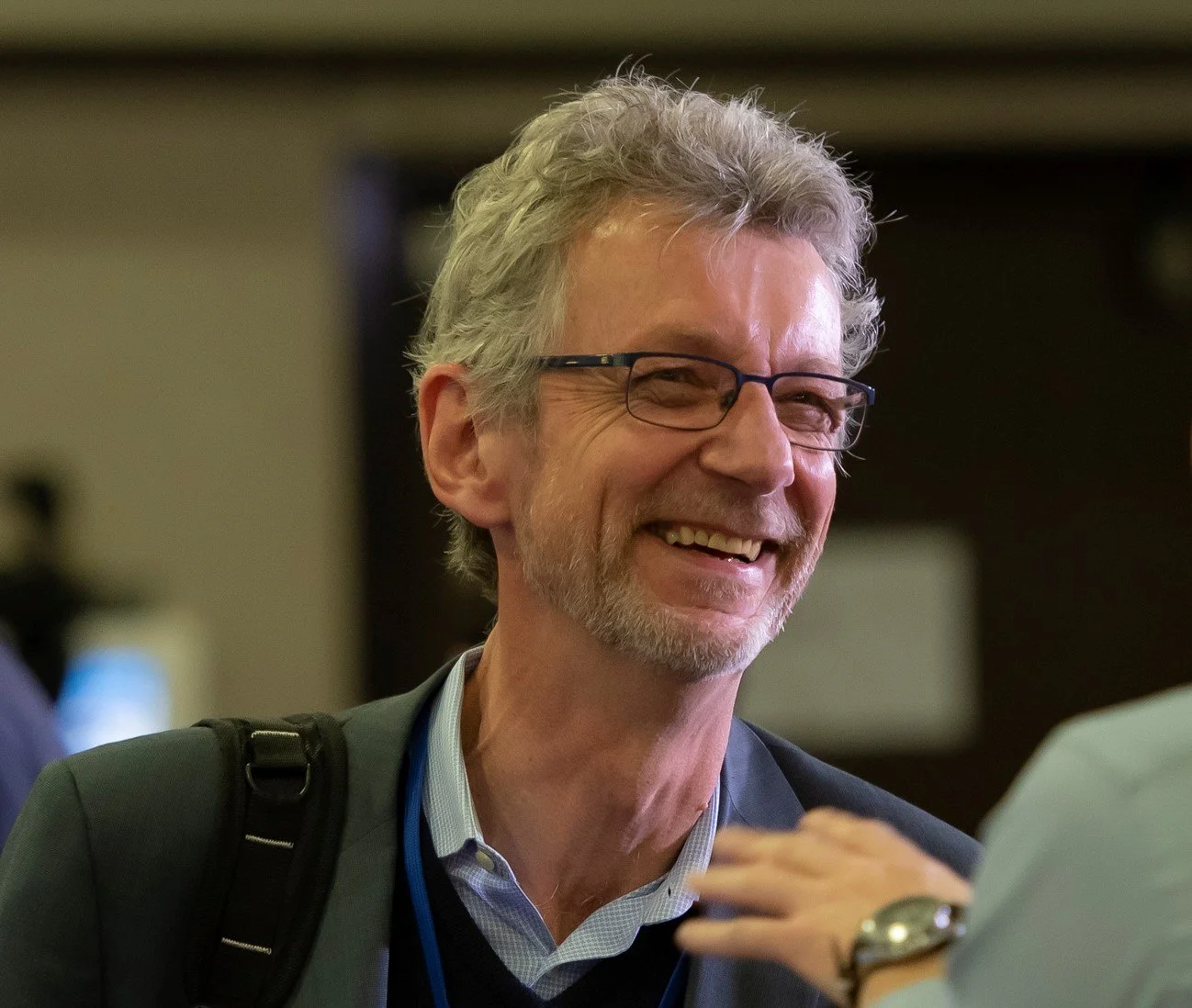 A smiling older man with glasses and gray hair wearing a suit and carrying a backpack, engaged in conversation.