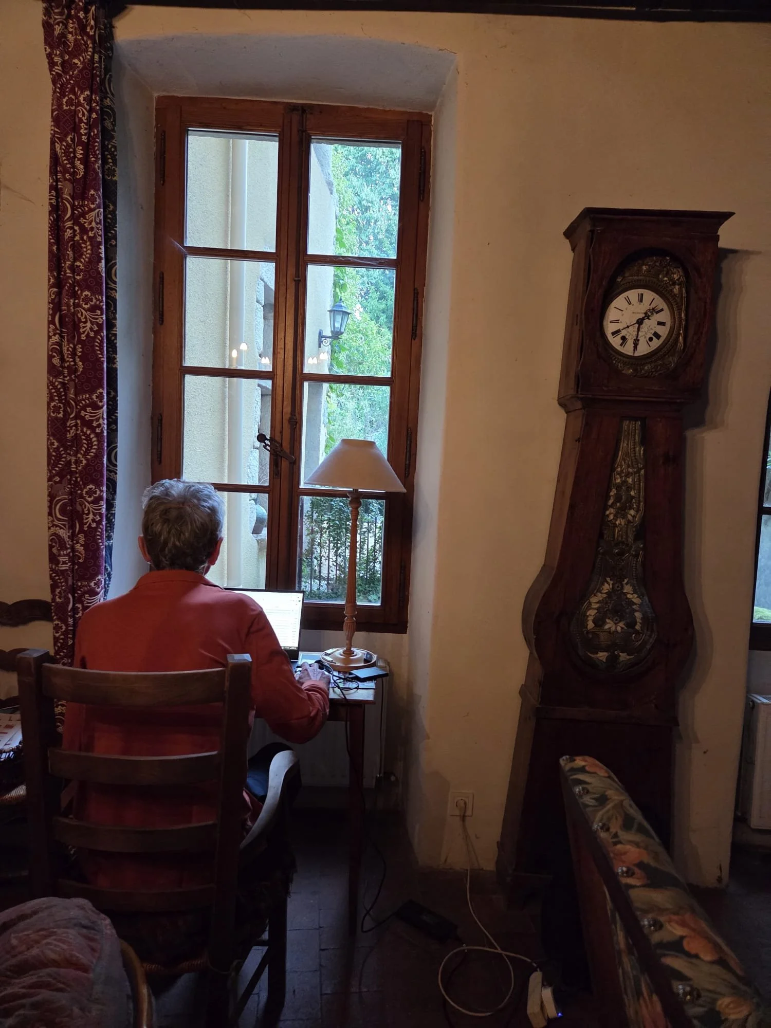 A person sitting at a small wooden table using a laptop, near a large window with wooden frames, outside trees and a street lamp are visible, a vintage wooden clock on the wall, a lamp on the table, and patterned curtains on the side.
