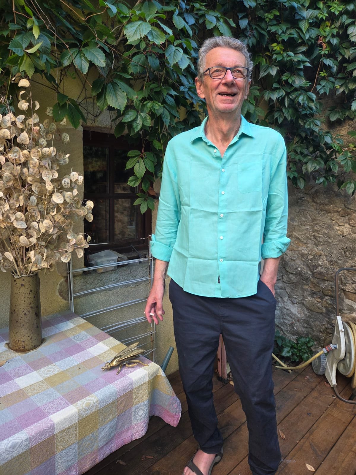 An elderly man wearing glasses, a light blue button-up shirt with sleeves rolled up, and dark pants, stands outdoors in front of a lush leafy background. He is smiling and has one hand in his pocket.